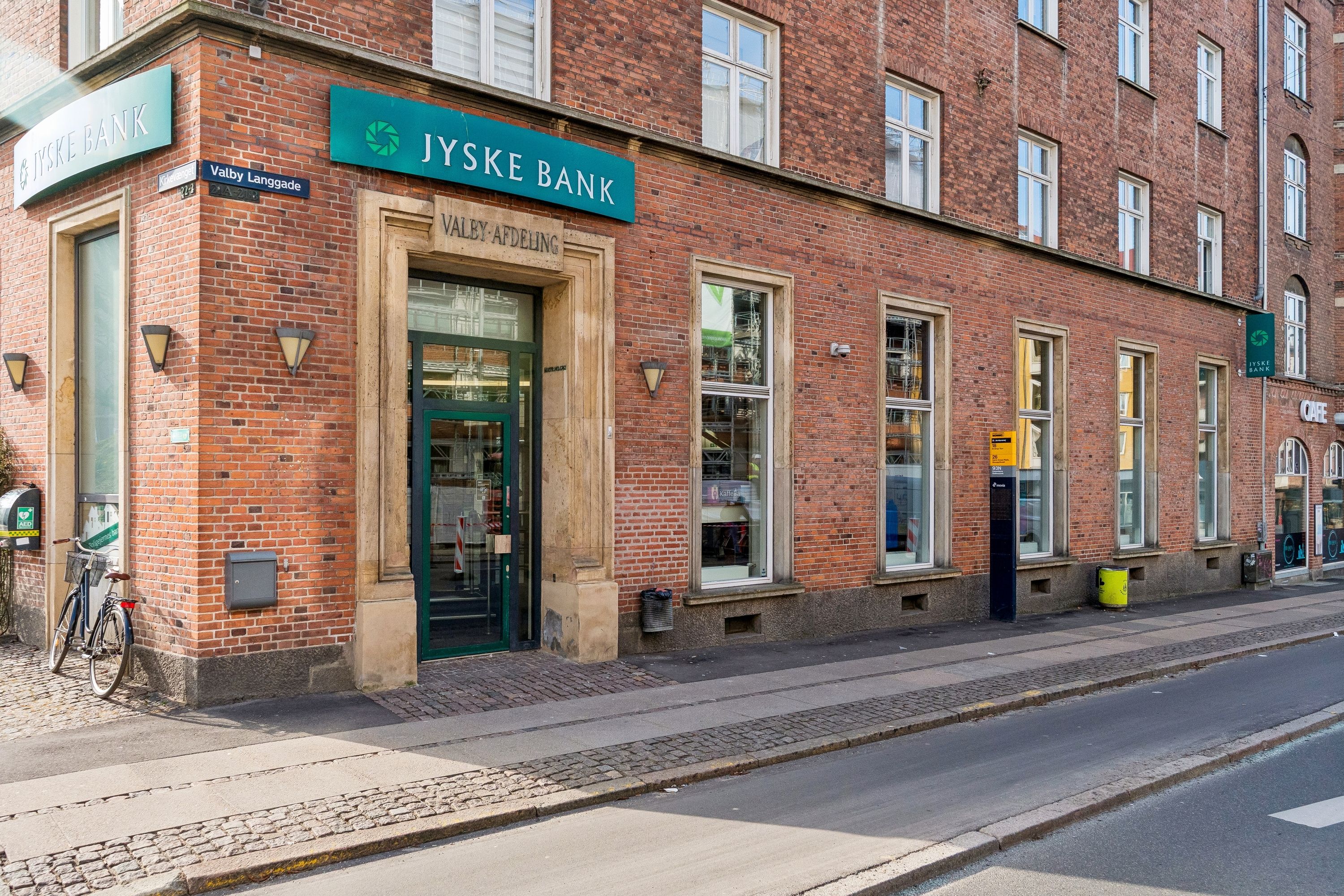 En rødstenebygning med Jyske Bank skilt og Valby Langgade 21. En cykel står foran døren og en gul skraldespand er placeret på fortauet.