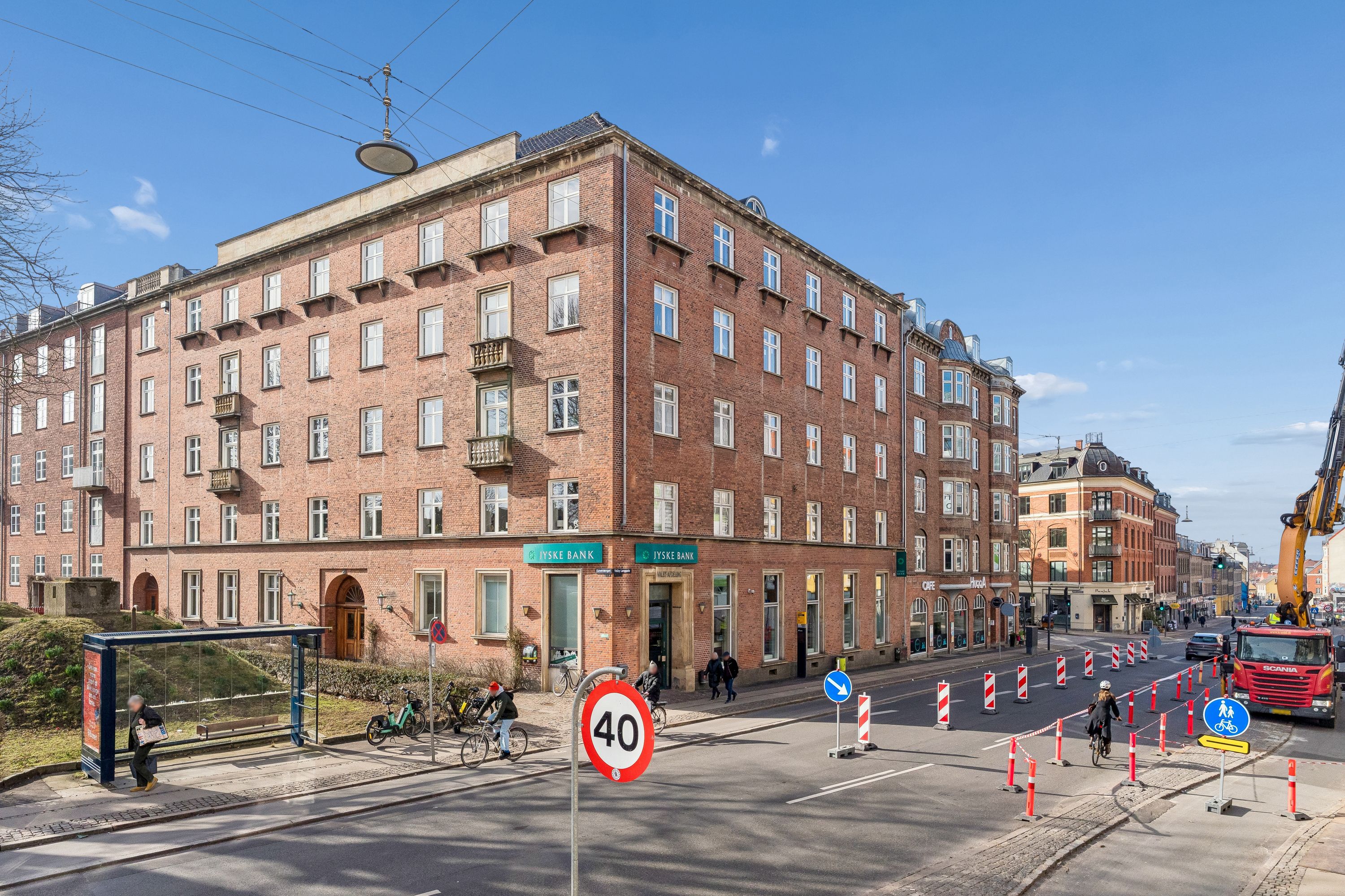 Kontorbyggeri i Valby med røde sten og lyseruder. Ydre 40 km/t skilt og trafikkegle. En person cykler forbi og en person læser avis på busstop.