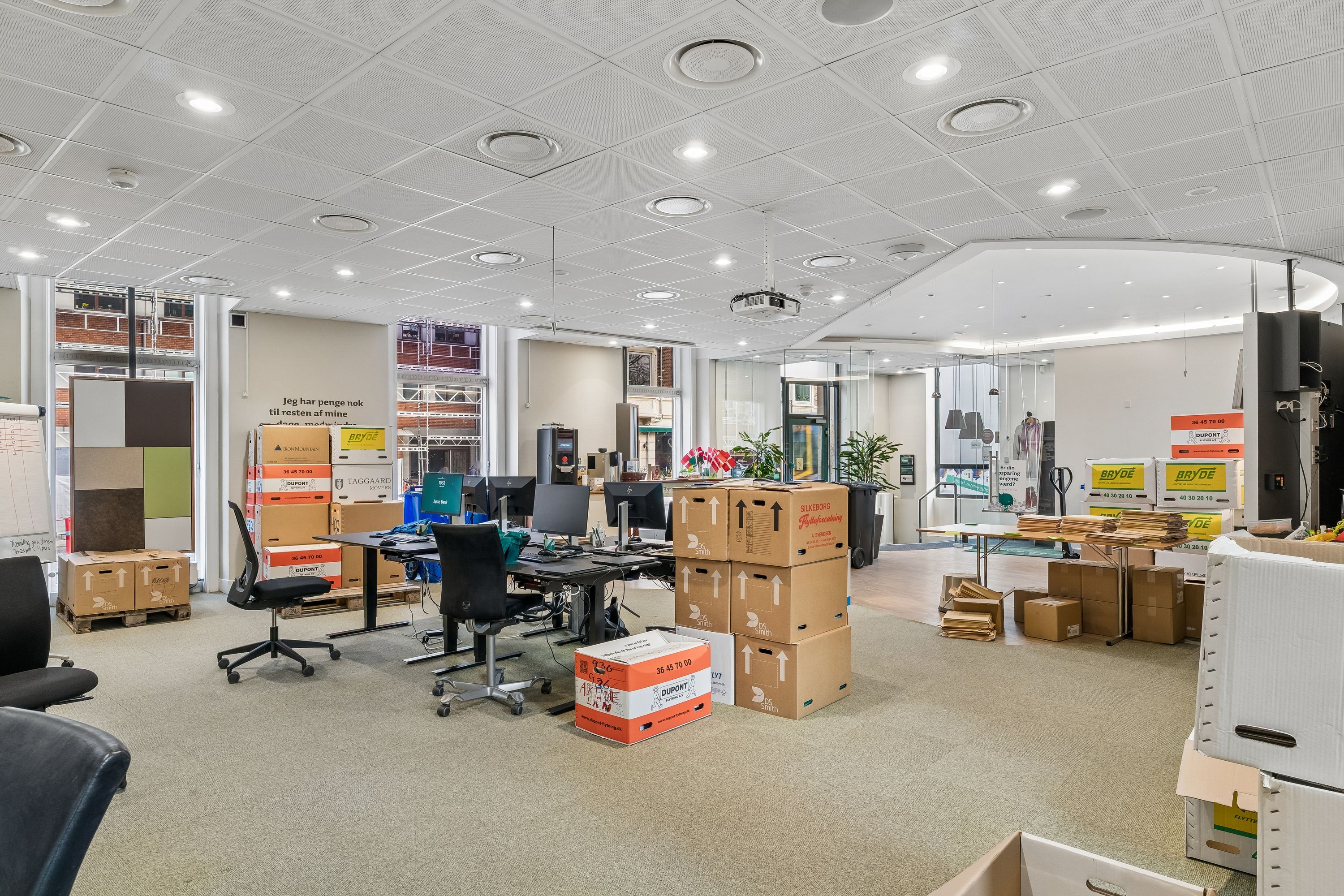 Kontor med skriveborde, stole, papkasser og computere. Lys gulvbelægning og lyse loftslampe. Planter og vinduer med udsigt til bygninger og gader.