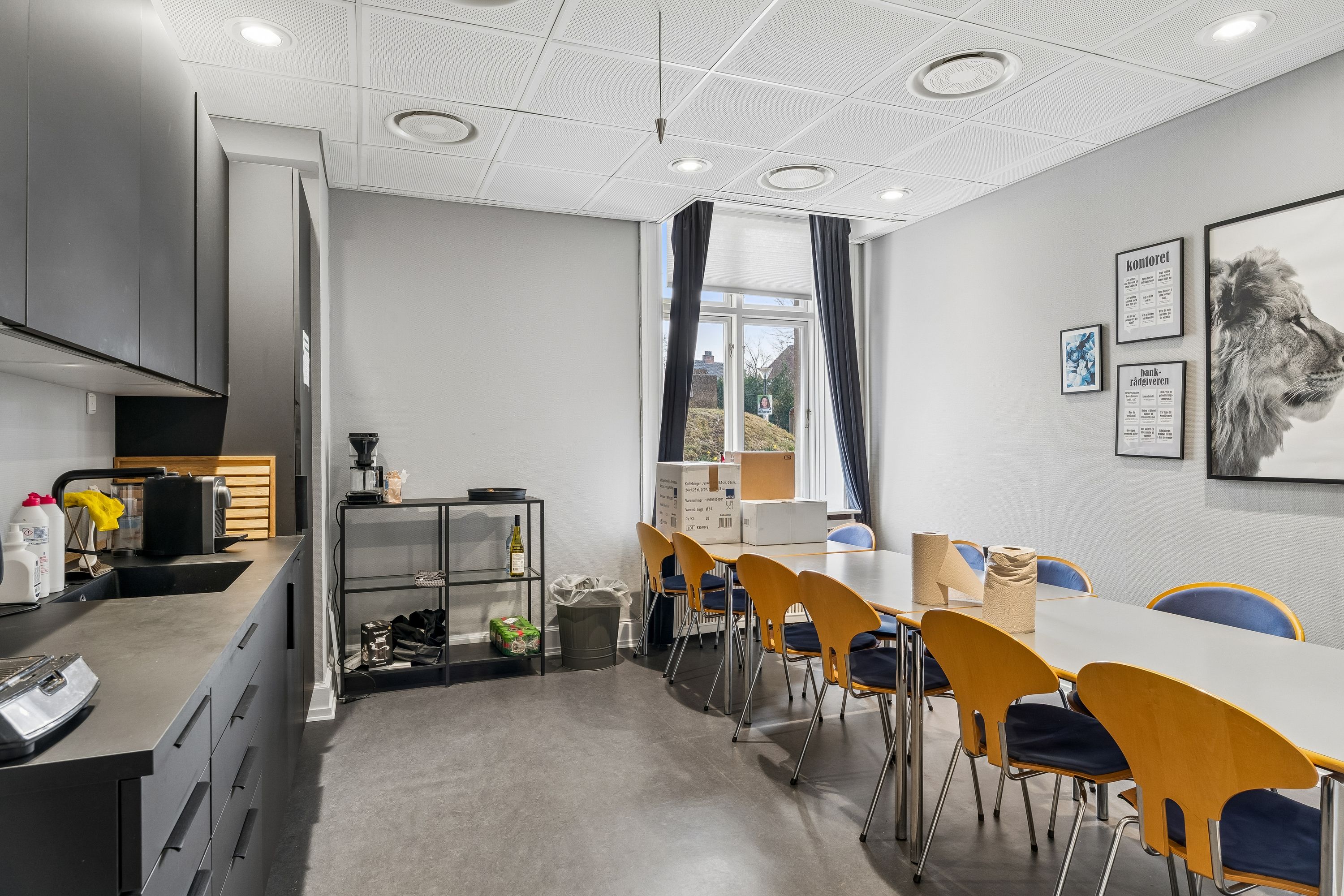 Kontor med lyse møbler og glasvinduer med gardiner. Væggene er hvide med kunstværker og plakater. Køkken med komfur, kaffemaskine, flasker og skabe. To lange borde med lysestole og servietter.