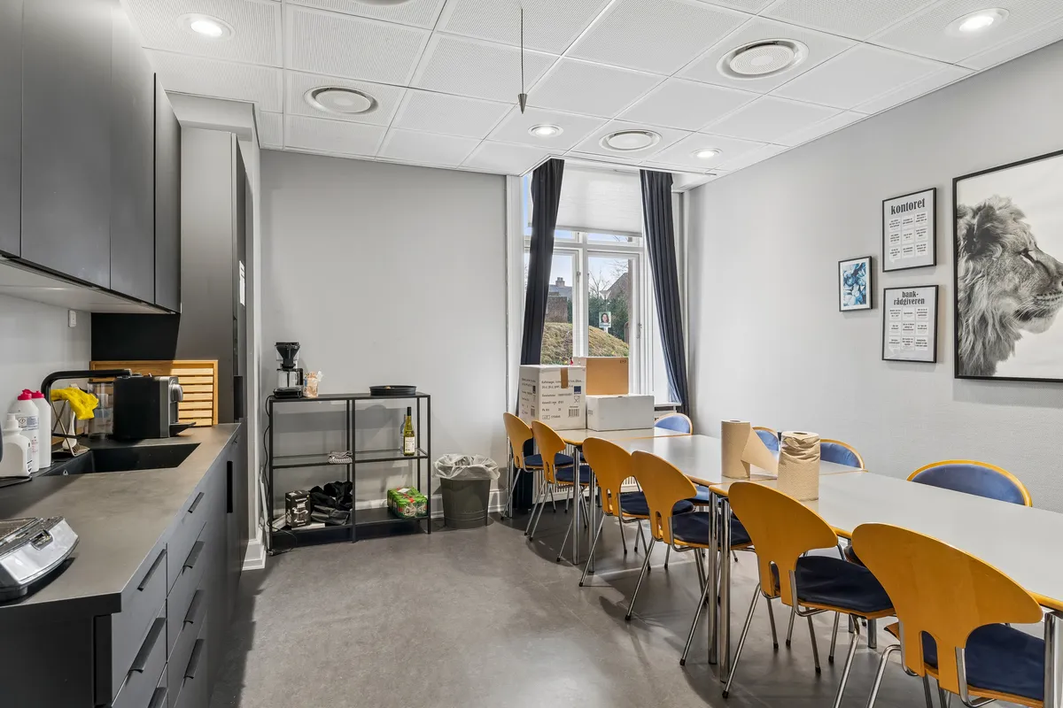 Kontor med lyse møbler og glasvinduer med gardiner. Væggene er hvide med kunstværker og plakater. Køkken med komfur, kaffemaskine, flasker og skabe. To lange borde med lysestole og servietter.