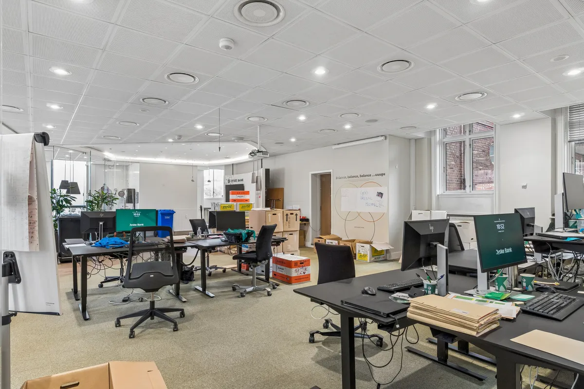 Kontorrum med to store skriveborde, flere mødestolhe, computere og to mødeborde med aviser og kuverter. Gulvet er tæppe, og der er flere kasser og kartoner i rummet