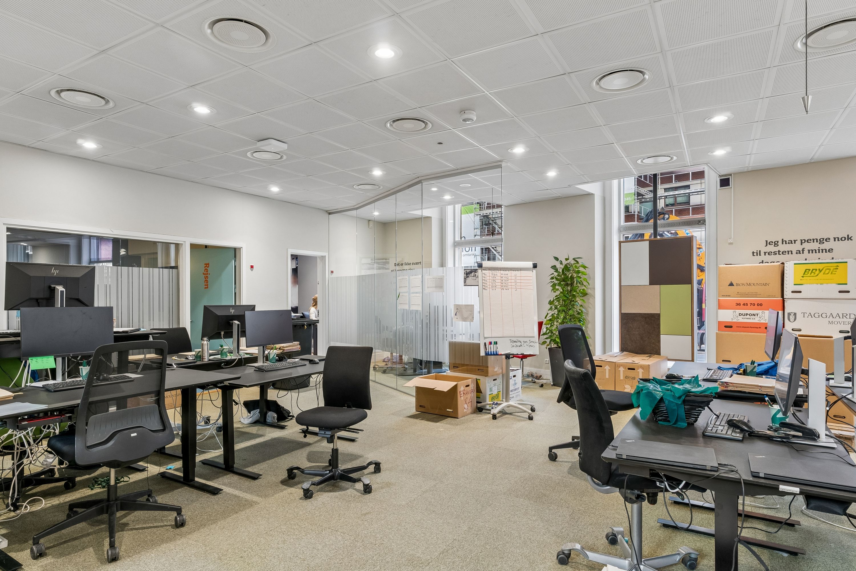 Kontor med skriveborde og stole, monitorer og tastaturer, forskellige kasser og bokse, plante og en glasdør.