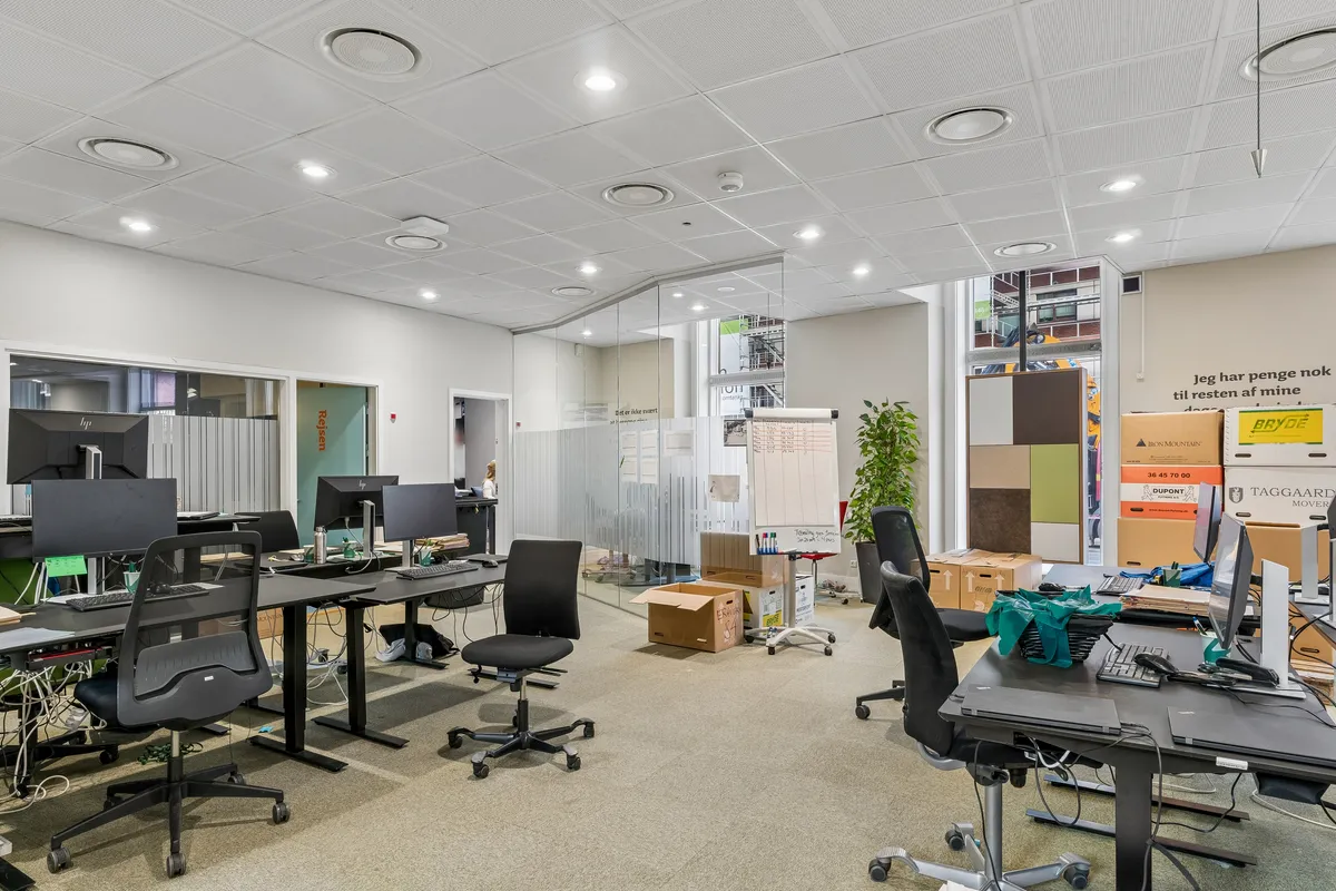 Kontor med skriveborde og stole, monitorer og tastaturer, forskellige kasser og bokse, plante og en glasdør.