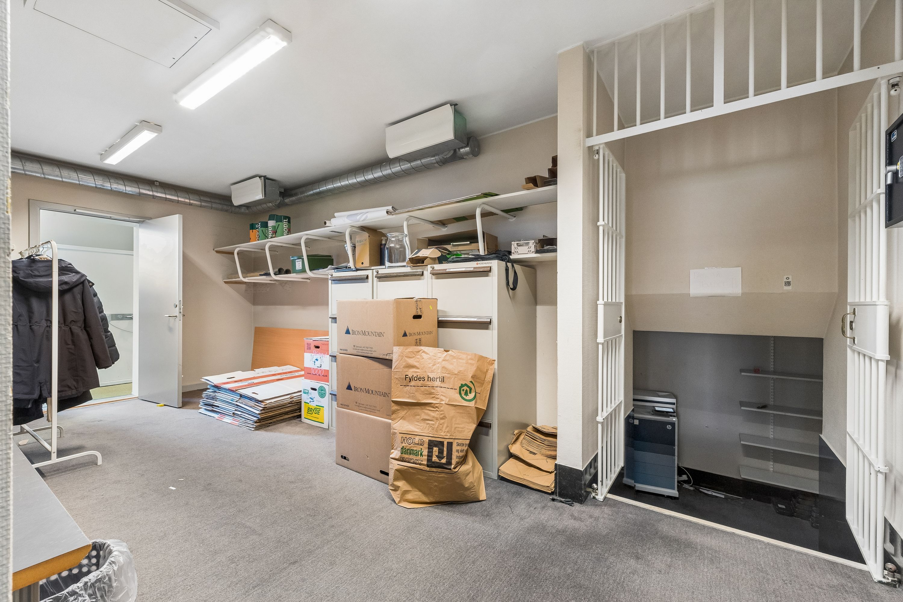 Kontorrum med store lagerkasser og papkasser. Mange hylder med diverse ting og høj gulvbeklædning. Værelse med møbler og vinduesdør. Luftkanaler og enheder på loftet.