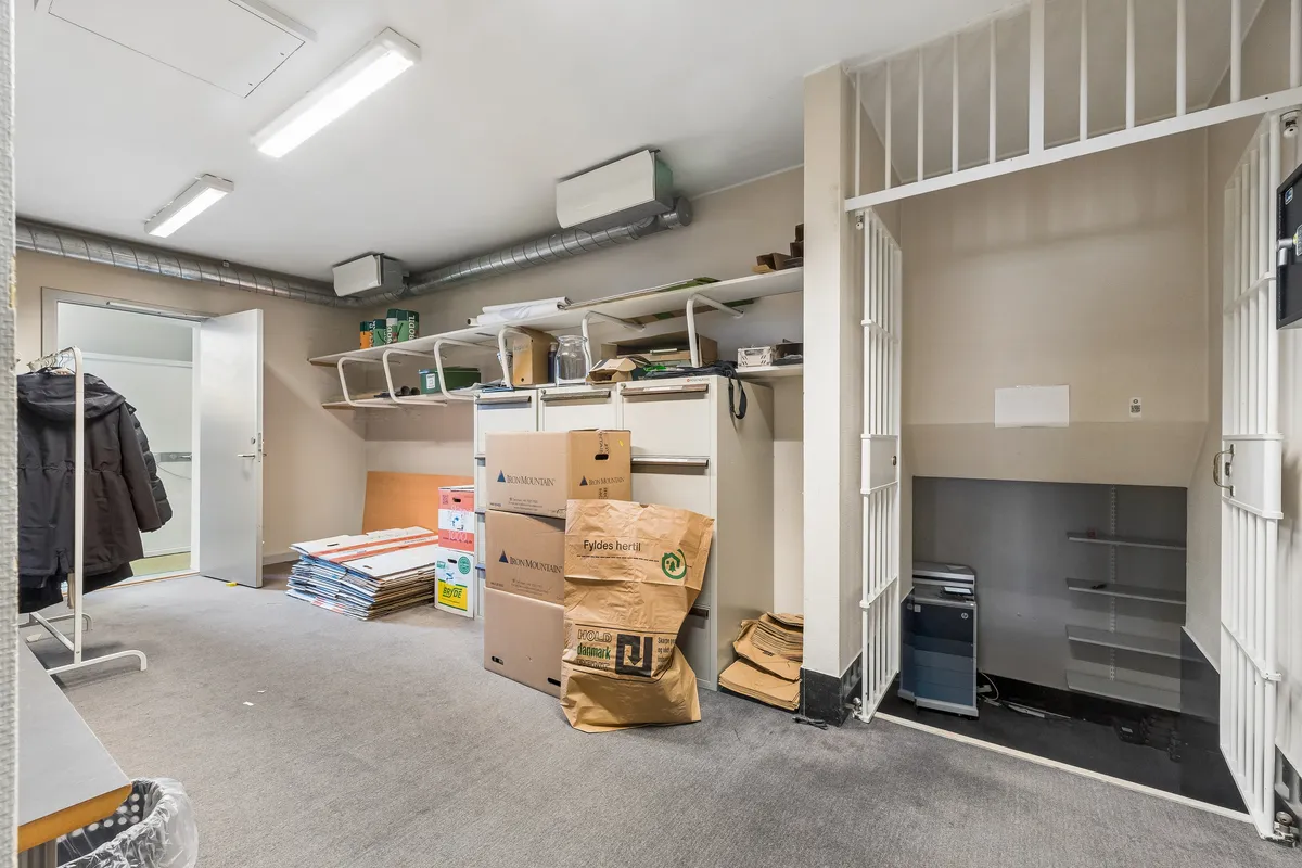 Kontorrum med store lagerkasser og papkasser. Mange hylder med diverse ting og høj gulvbeklædning. Værelse med møbler og vinduesdør. Luftkanaler og enheder på loftet.