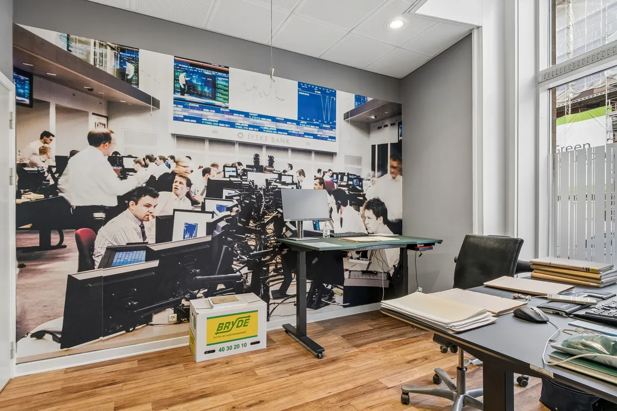 Kontor med lysegul væg og møbler med skrivebord, stol, og monitor, udstyr som bøger og tastature. Vægbillede med arbejdere.