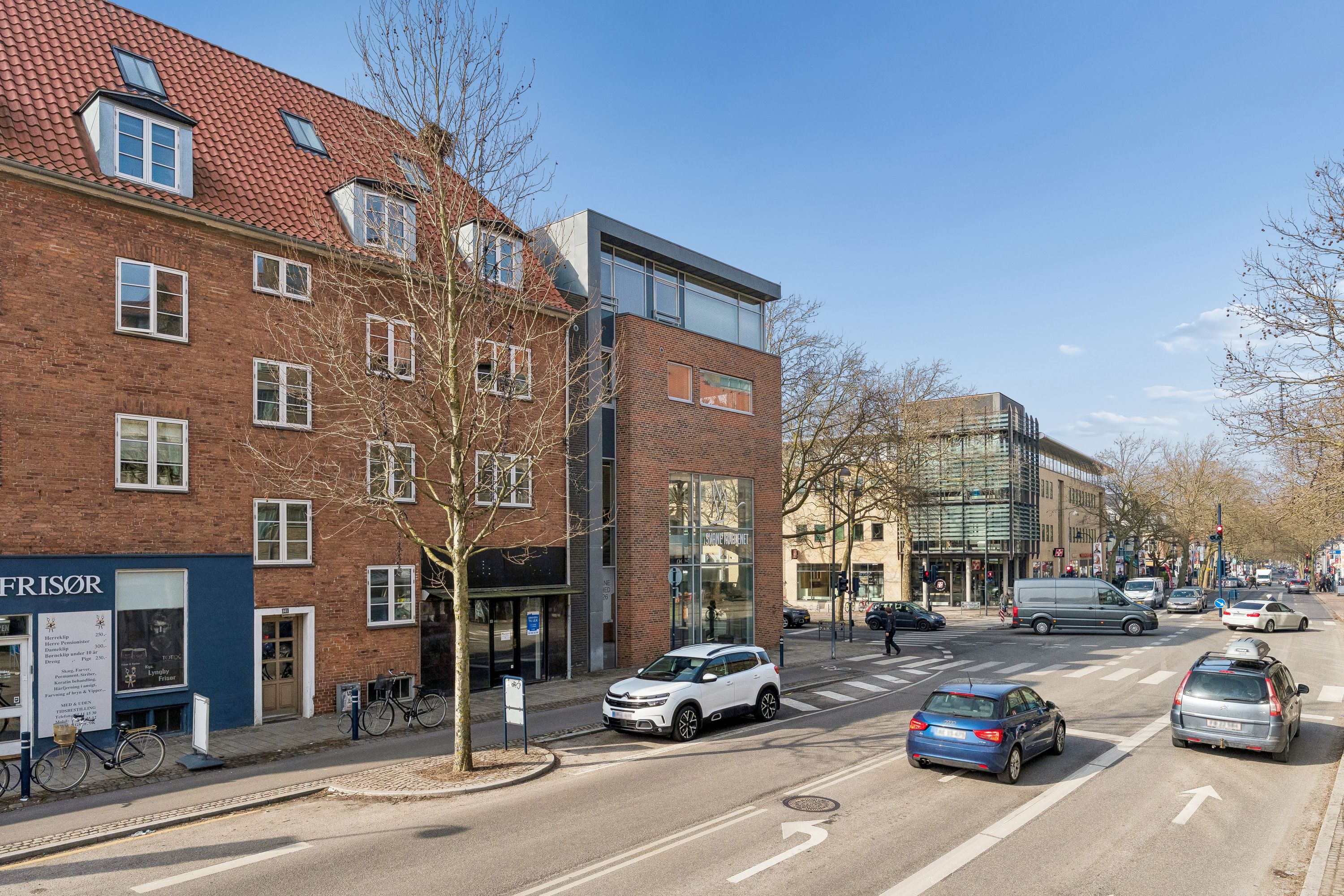Vejkryds med biler i Kongens Lyngby, der kører på Lyngby Hovedgade, med fortov, cykelstel, og rødt lys. I baggrunden er der store bygninger og en rødt tag på et lejlighedshus.