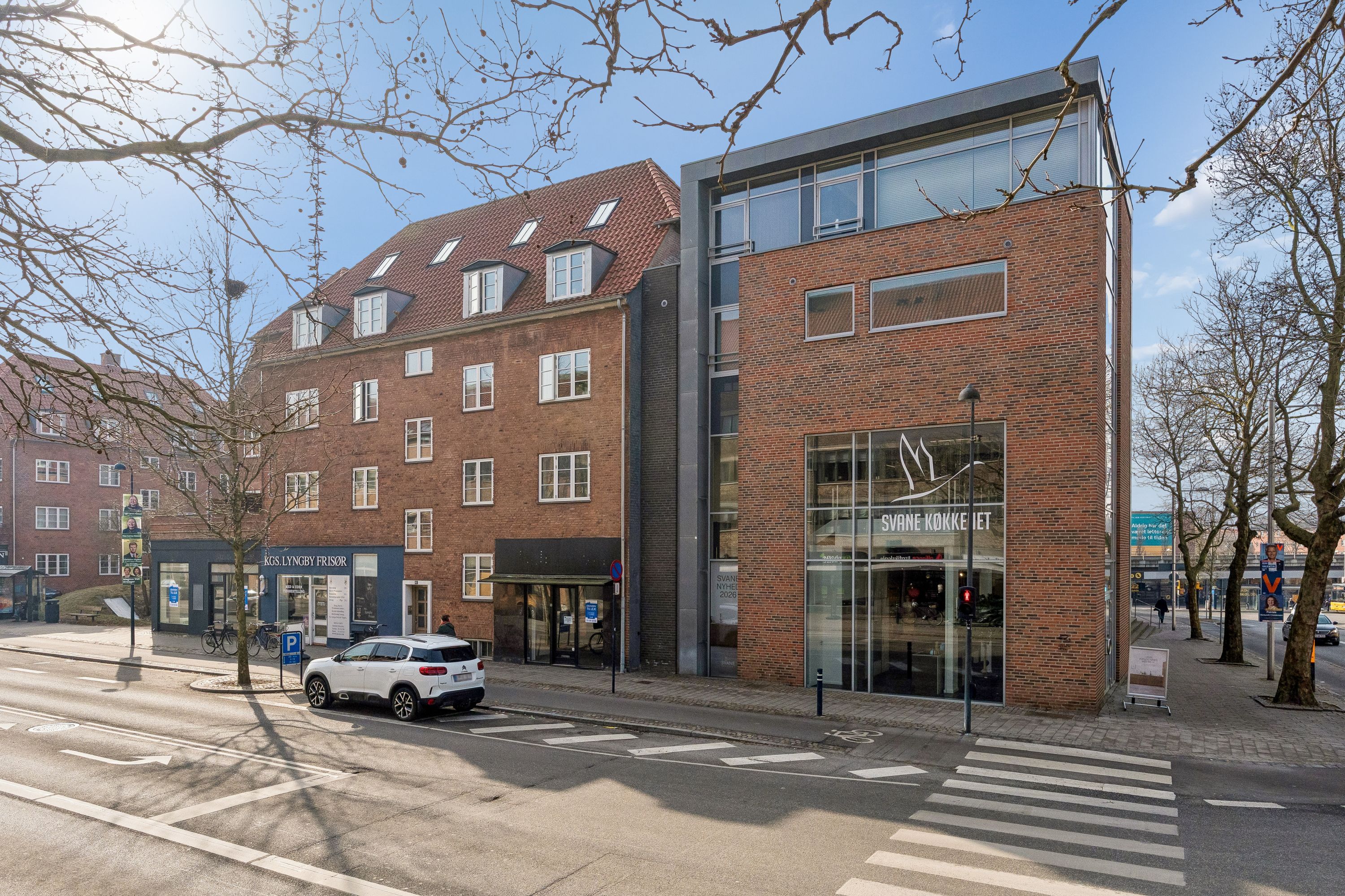 En hvid SUV parkeret på gågaden, med butiksvinduer og en stoplygte i baggrunden. Et større butikslokal med glasdøre og teksten Svane Nyheder 2026.