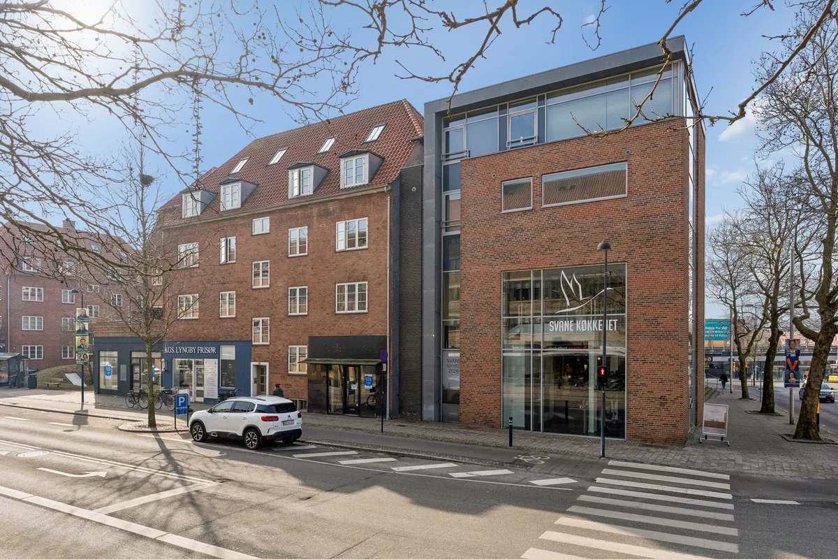 En hvid SUV parkeret på gågaden, med butiksvinduer og en stoplygte i baggrunden. Et større butikslokal med glasdøre og teksten Svane Nyheder 2026.