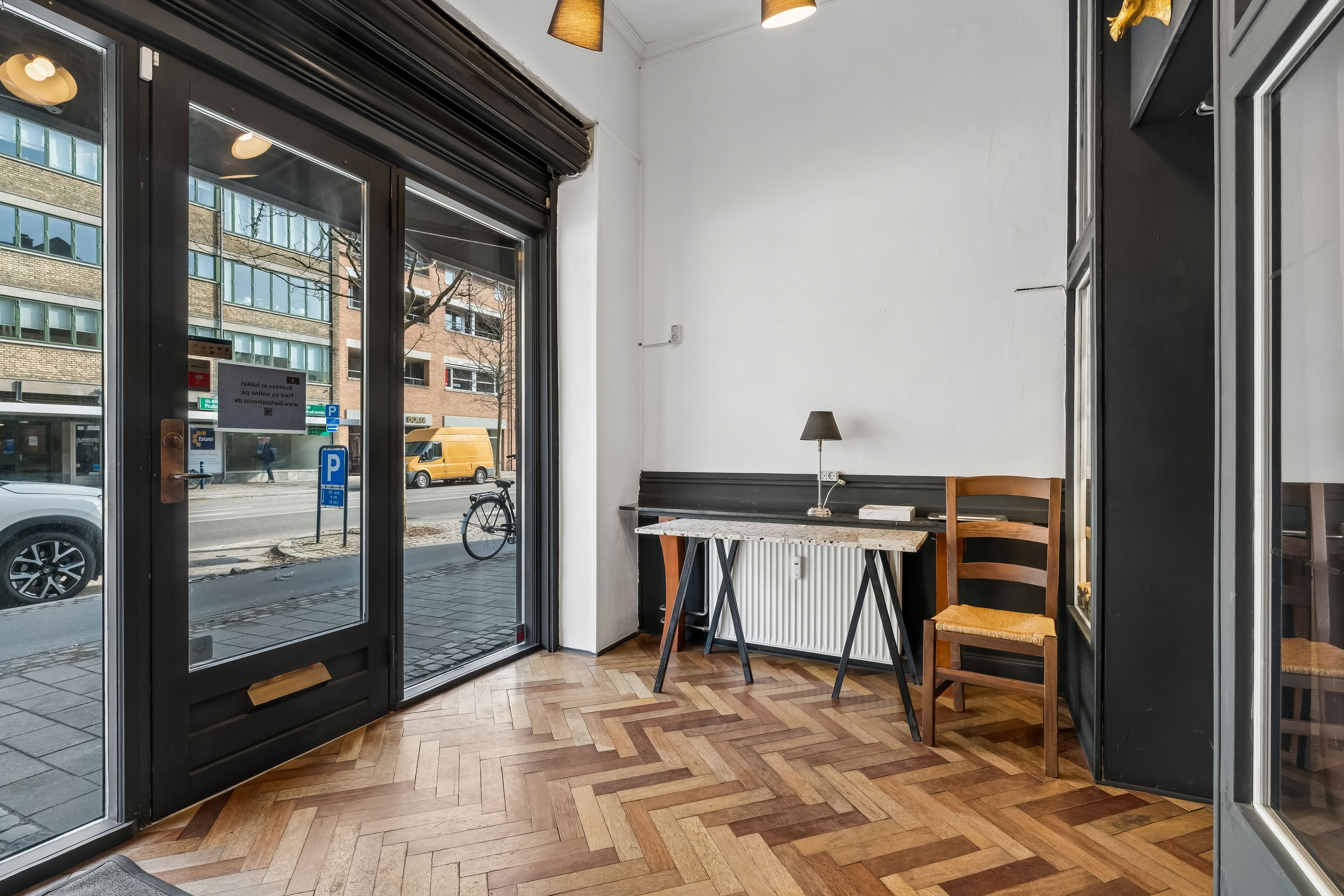 Indenfor er en ren stue med trægulv og højloft. Der er en bord og en stol med en lampe. Dørene er glas med mørke kanterne. Der er et køretøj og en cykel parkeret udenfor.