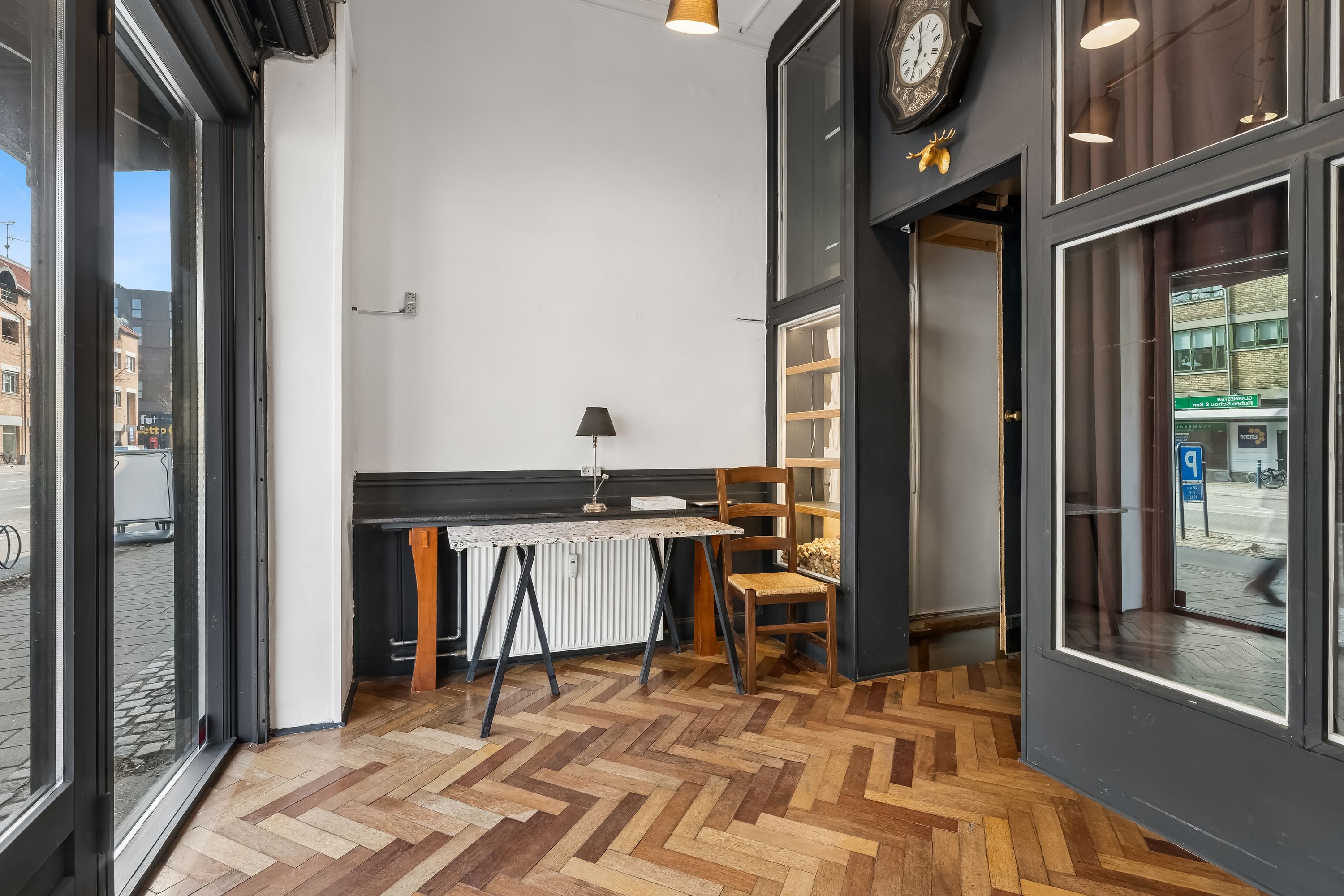 Her er en elegant stue med trægulv, lyse vinduer og møbler. Et køkkenbord med lampe og en stol. Et ur og et fasanhoved på væggen. Et skrivebord med lysekrone på loftet.