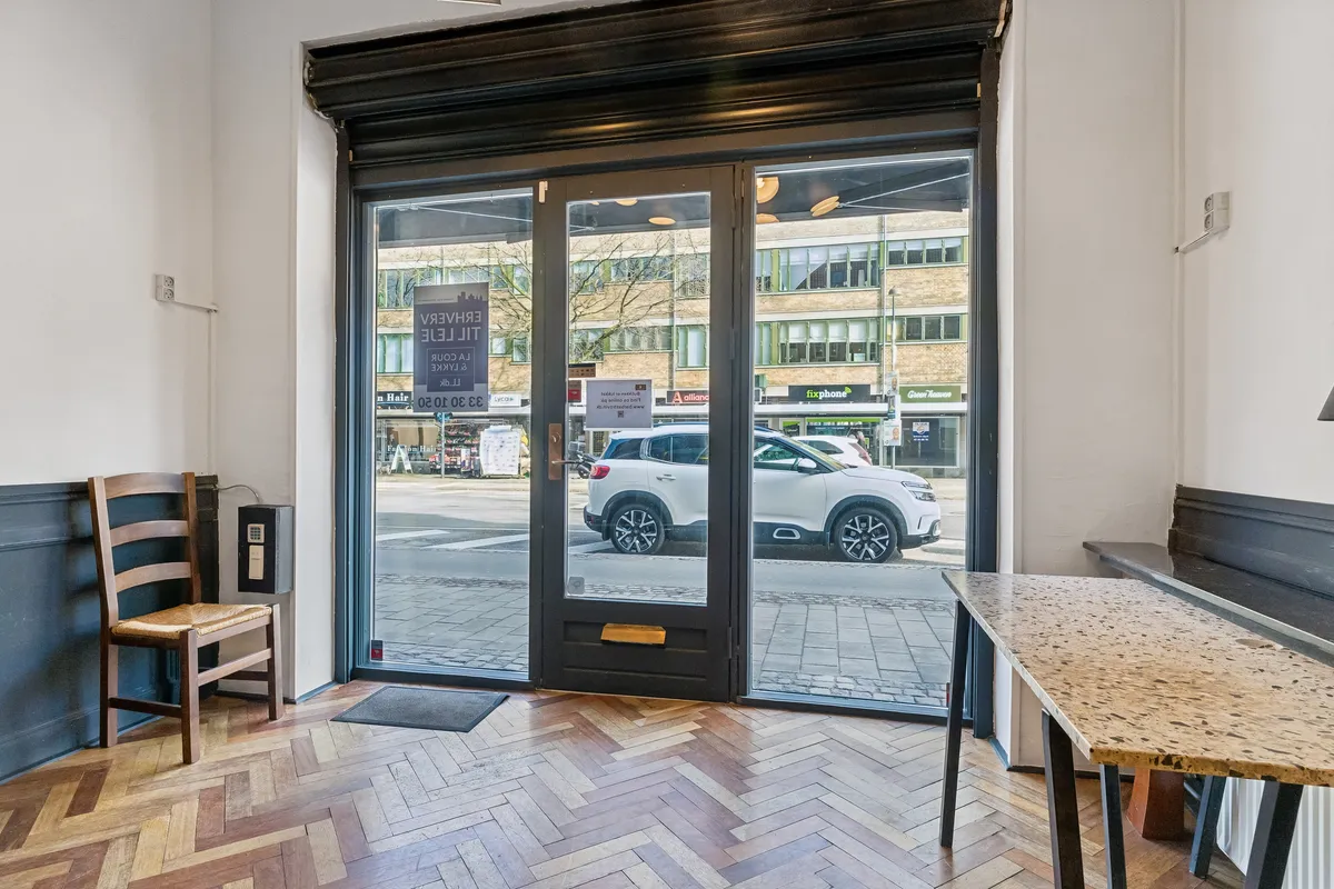 Butik til leje på Lyngby Hovedgade 84A, st. 1, 2800 Kongens Lyngby. Store glasdøre med indskrift og stol til gæster. Moderne interiør med betonbord og højt gulv