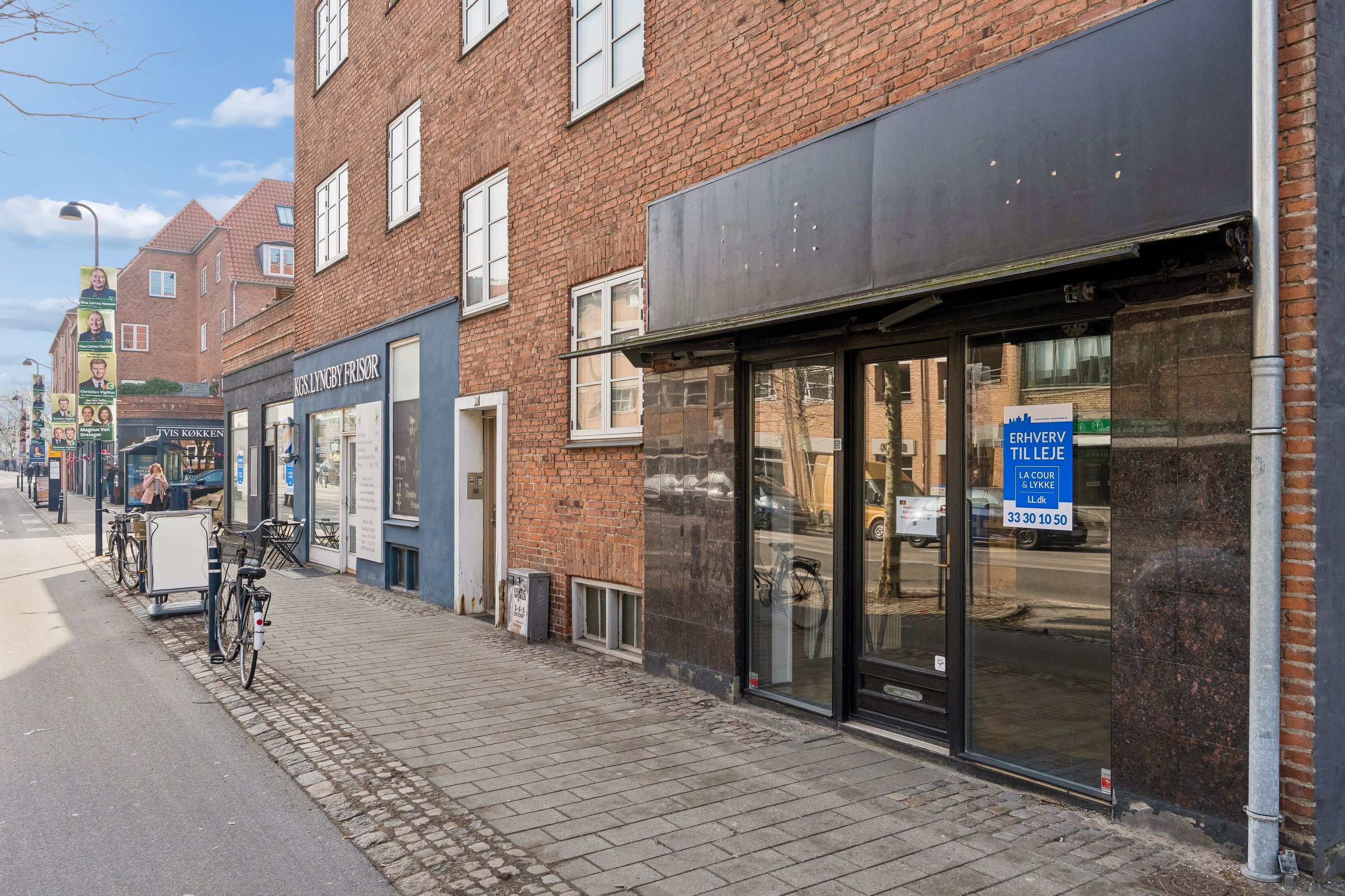 Foto af en stenbrolægget gade med butiksruder og skilte samt to parkerede cykler og en søjle med annoncer i Kongens Lyngby.