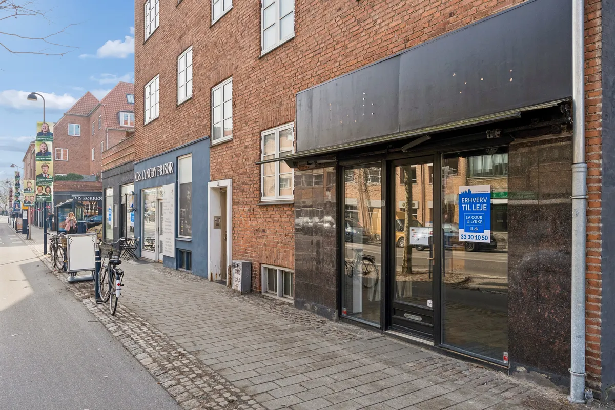 Foto af en stenbrolægget gade med butiksruder og skilte samt to parkerede cykler og en søjle med annoncer i Kongens Lyngby.