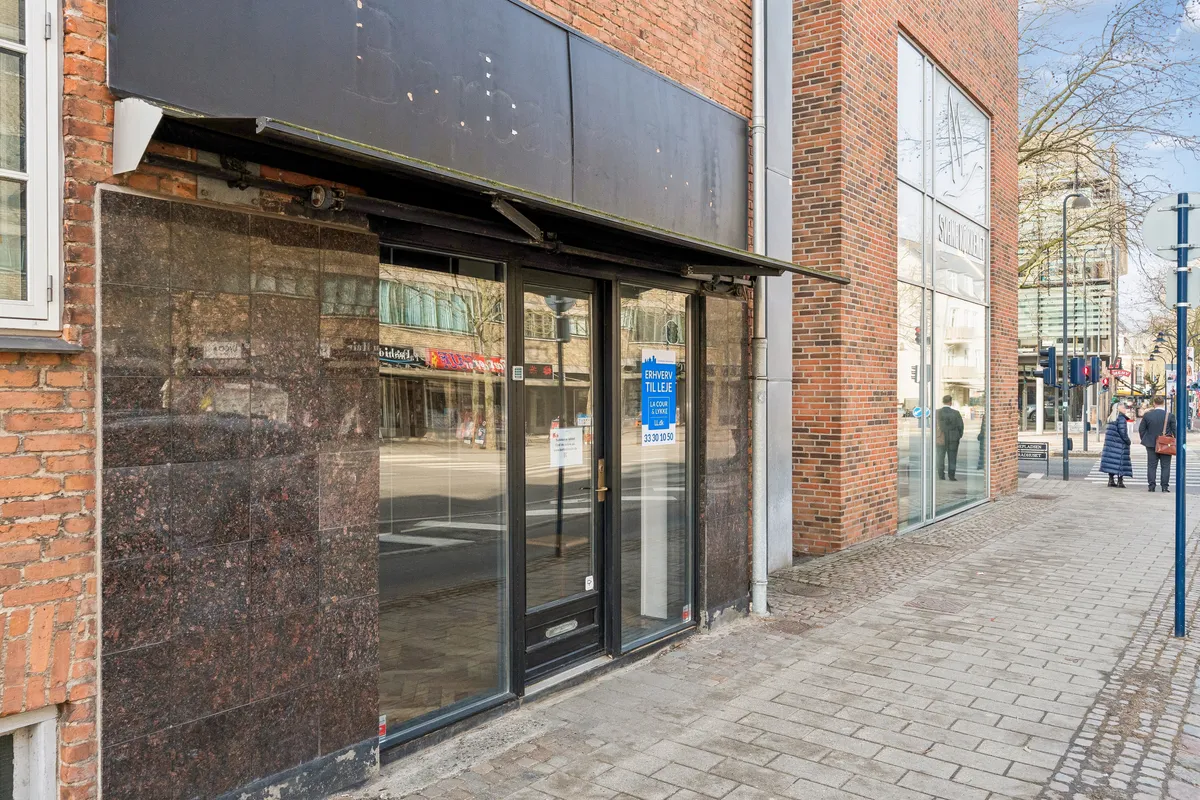 Lejlighed til leje i Kongens Lyngby med butiksvinduer og glasdøre, søjler og en fortovskant. To personer krydser gaden og en søjle med skilte står på fortovet.