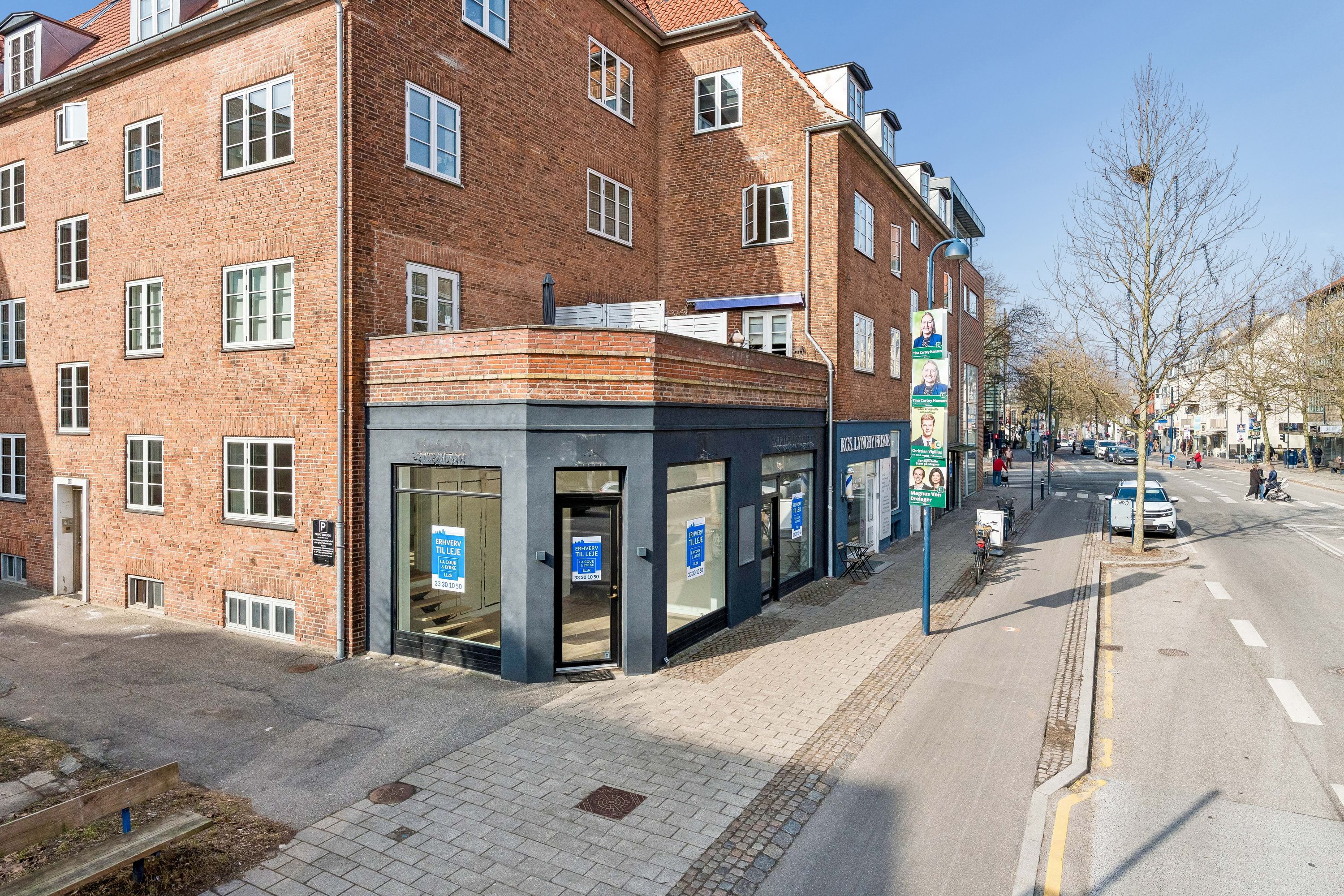 Butik til leje på Lyngby Hovedgade. Lejlighedsbygning med glasvinduer og annoncer på facaden. Parkerede biler og cykler langs siden af gaden. Mennesker og træer langs vejen.
