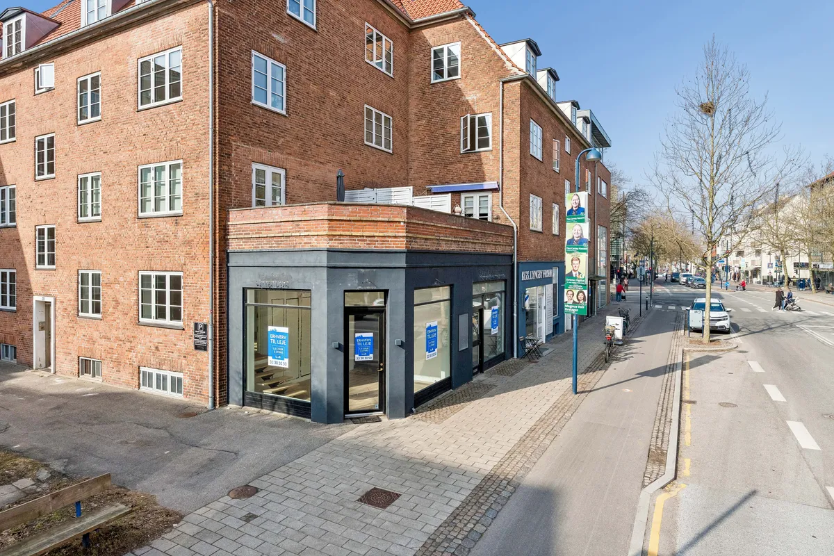 Butik til leje på Lyngby Hovedgade. Lejlighedsbygning med glasvinduer og annoncer på facaden. Parkerede biler og cykler langs siden af gaden. Mennesker og træer langs vejen.