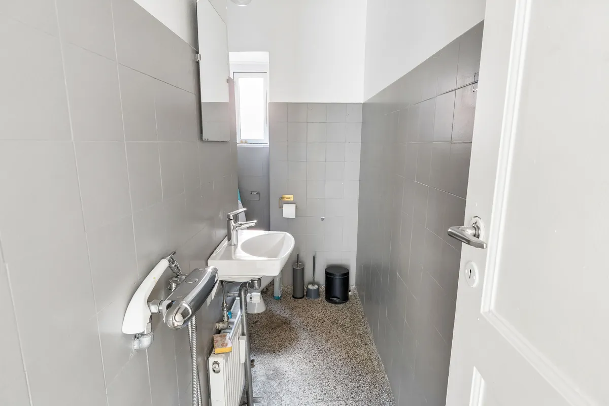 Badeværelse med hvid stålbadeværelsesudsmykning, hvidt keramikvask og hånddusch med metalhåndtag, gulvvarme og toilet på hjørne.