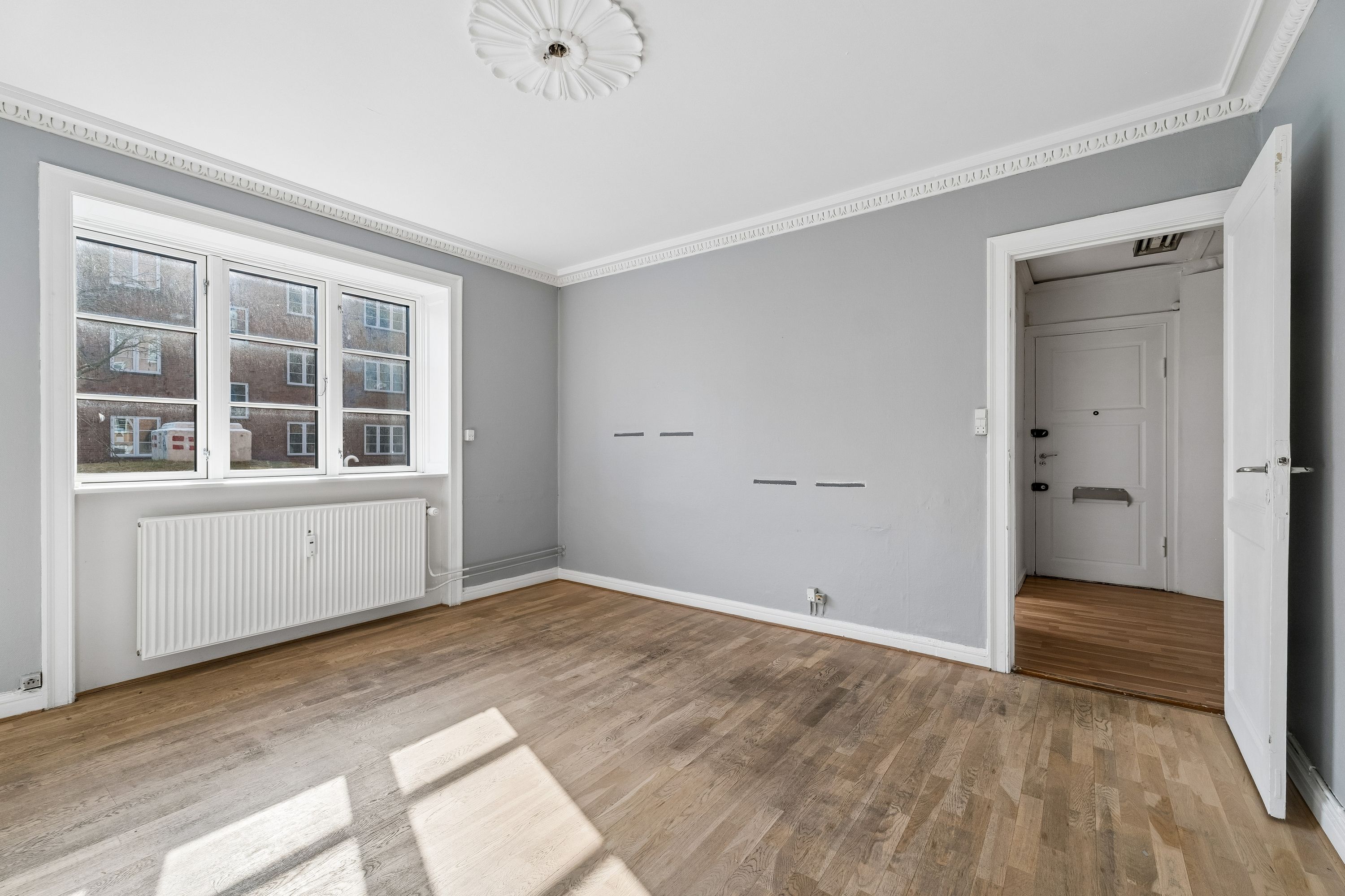 Store lyse stue med trægulv og hvide vægge. Vindue med refleksioner. Lejligheden har radiator og dør til et andet rum
