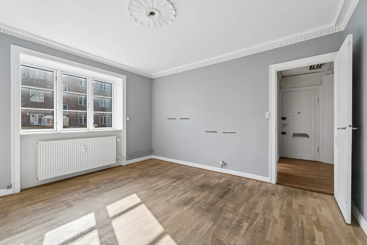 Store lyse stue med trægulv og hvide vægge. Vindue med refleksioner. Lejligheden har radiator og dør til et andet rum