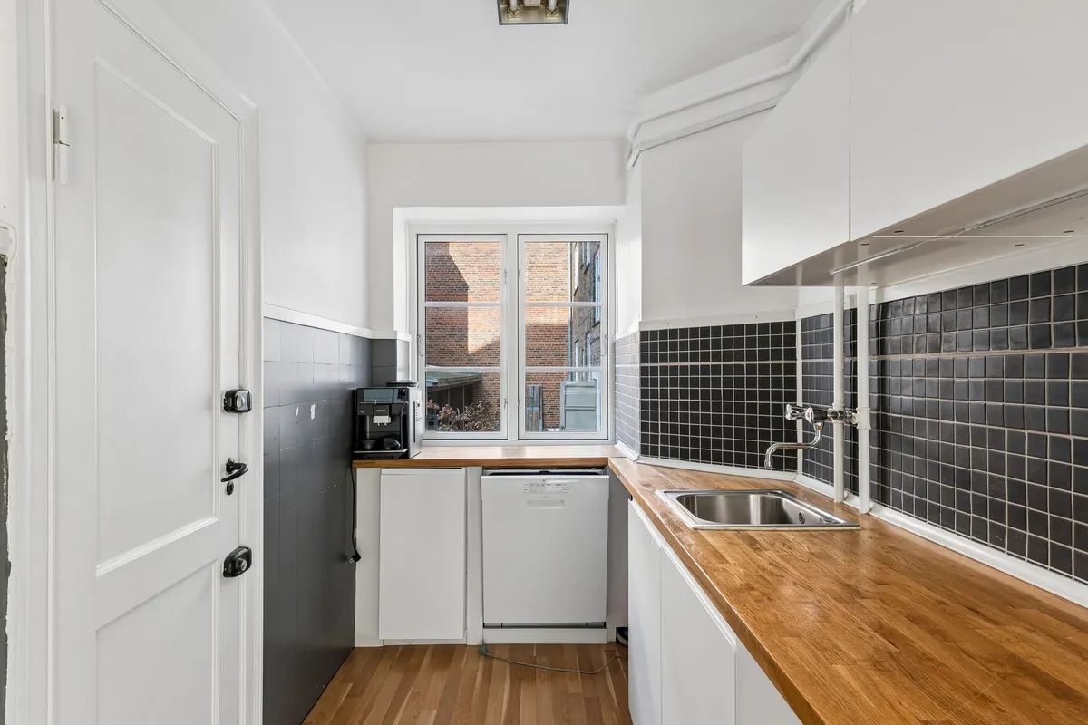 Minimalistisk køkken med hvide vægge og trægulv, et vindue med dobbeltdøre og udsigt til gården, en hvid opvaskemaskine og træbenk med håndvask og en sølvfarvet vandhane