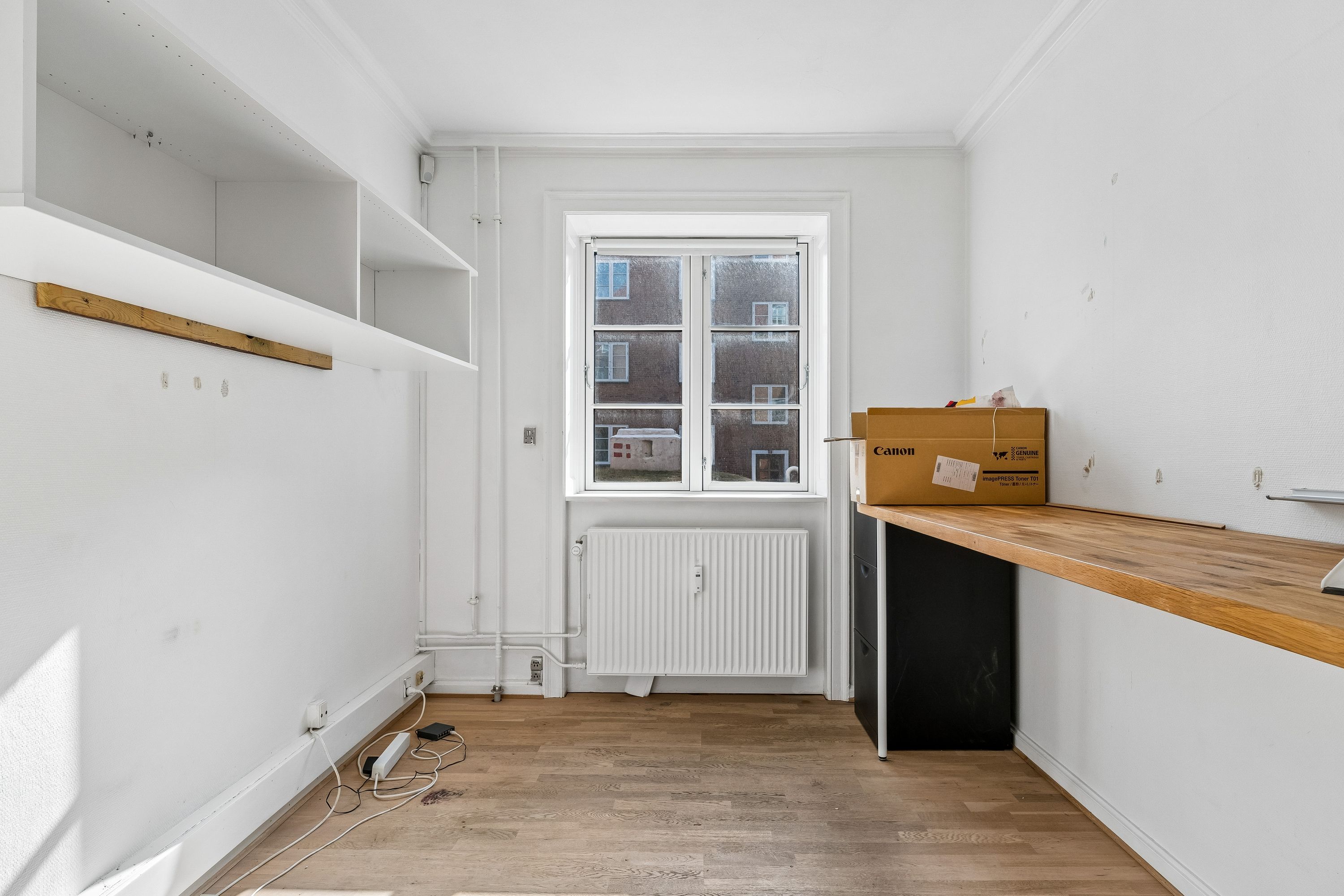 Tømt rum med lysegult gulv og hvide vægge. Højt kontorbord og bokse på gulvet. Vindue med udsigt til andre bygninger. Højt hængende hylde med tomme hylder.