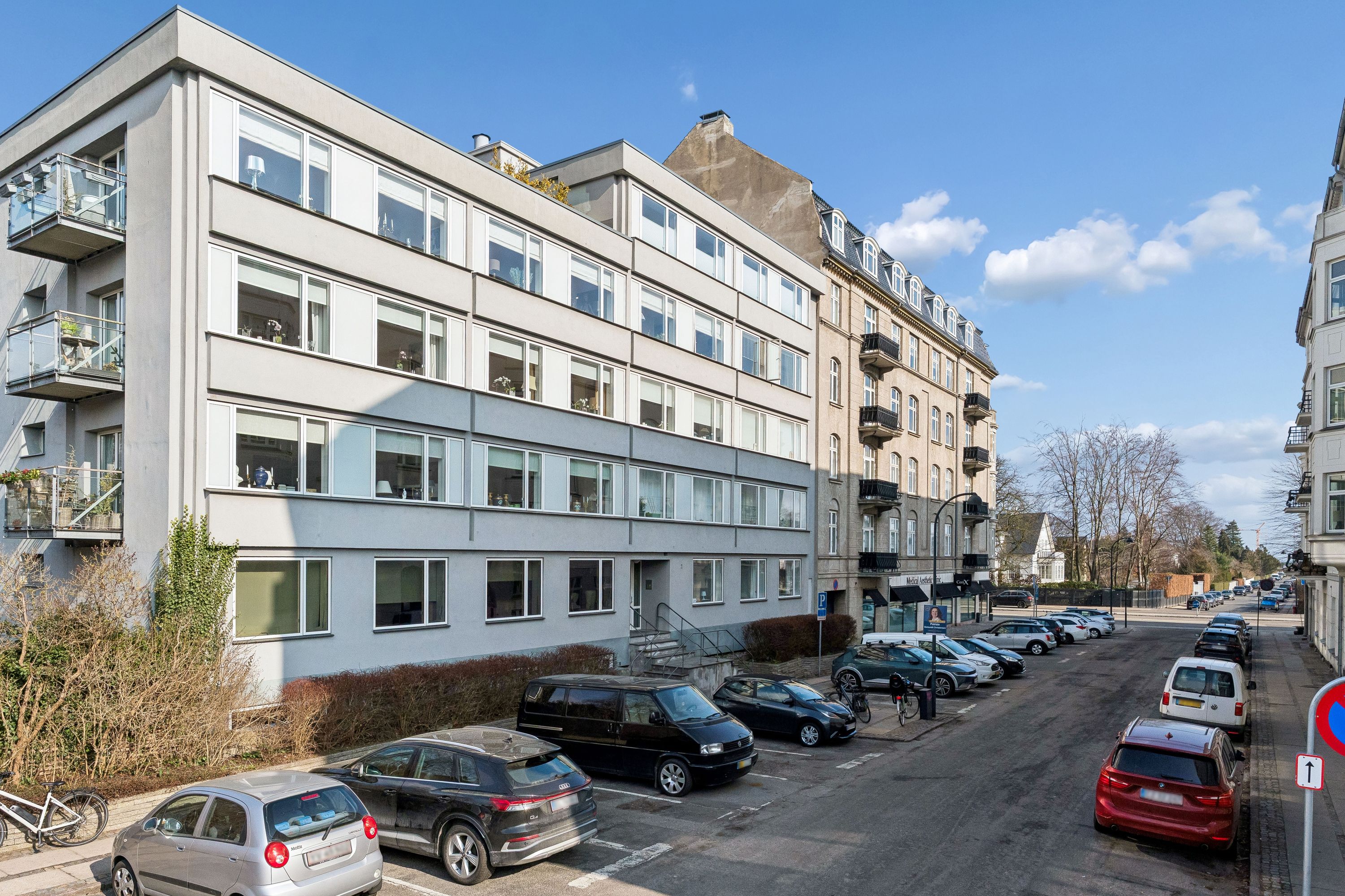 Kontorbyggeri med parkerede biler og cykler samt huse med balkoner i baggrunden