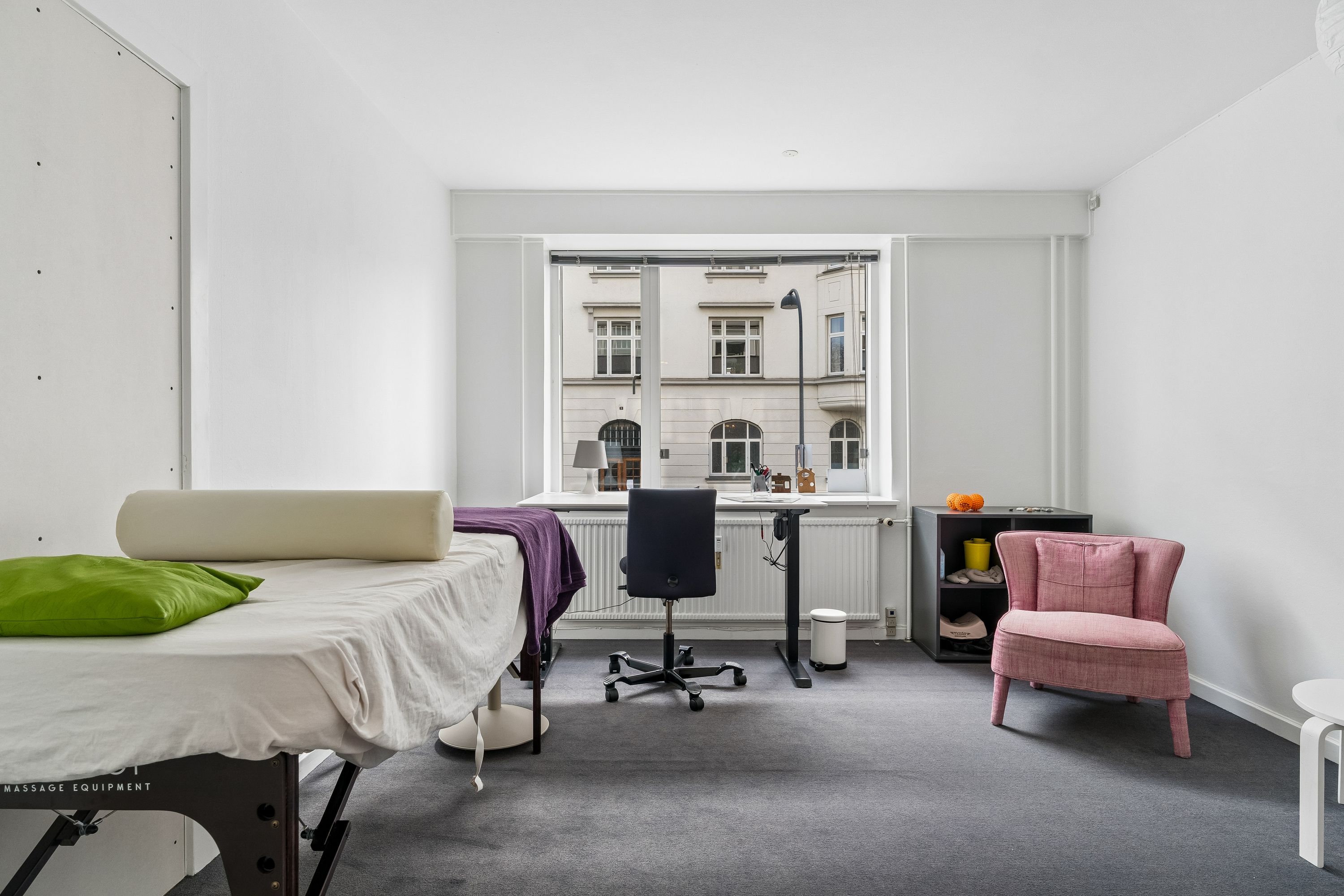 Moderne kontor med massagebord, skrivebord, stol og pink lænestol, lyserødt vindue, hvidt gulv, hvide vægge, gulvvarme og udsigt til Aurehøjvej