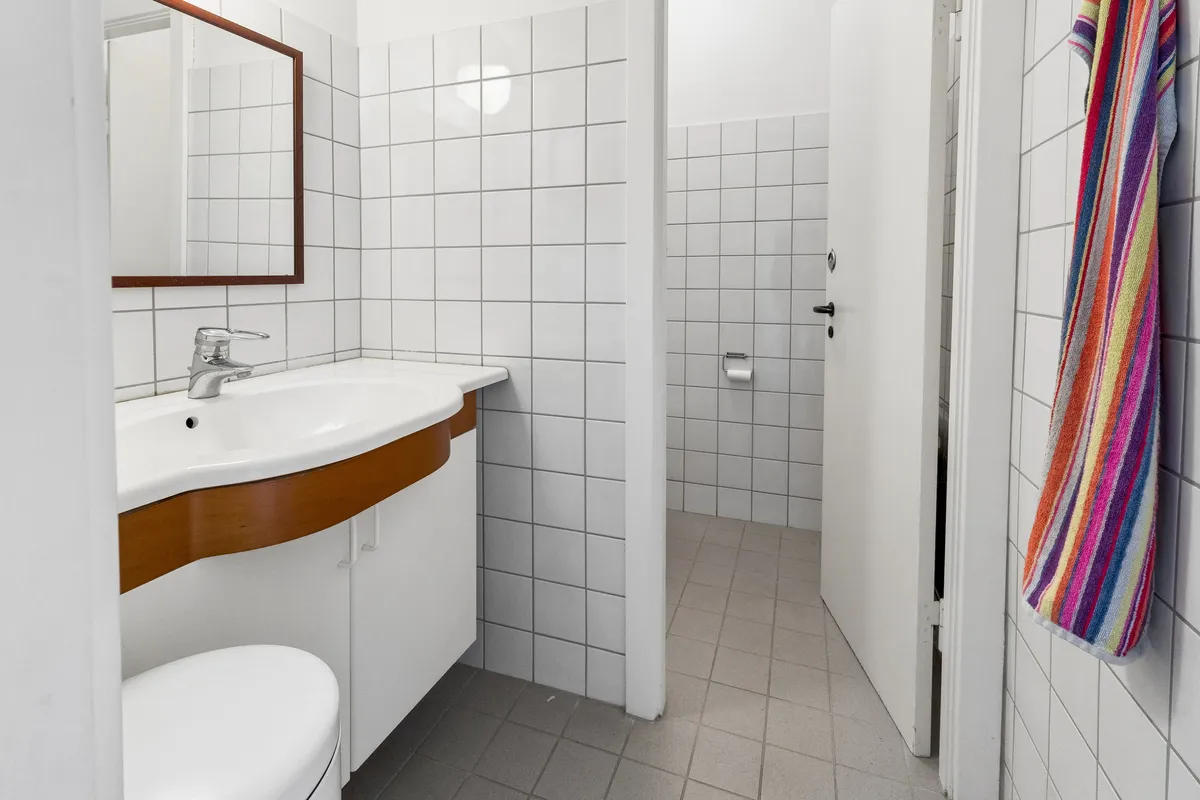 Ny, ren og pænt indrettet badeværelse med hvidt keramik og lysegrå gulvfliser samt en farvestrålende håndklæde på hængehylde