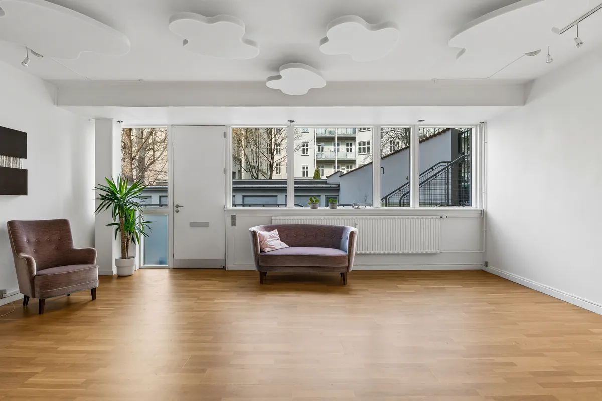 Kontor til leje i Aurehøjvej 2, st. th, 2900 Hellerup. To sofaleve, plante og hvide vægge. Moderne trægulv og vinduer med udsigt til gaden og et hus med trappe.