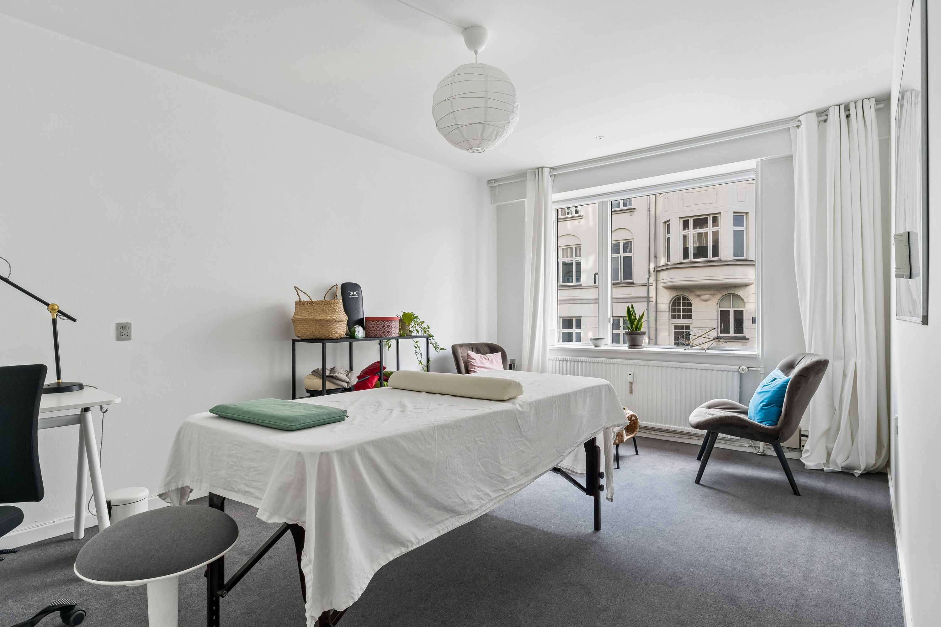 Et lyse og pænt kontorlokal med hvidt gulv, et træbord, et højt stol, tæpper og en massagebord med hvidt linne og flere tæpper og puder