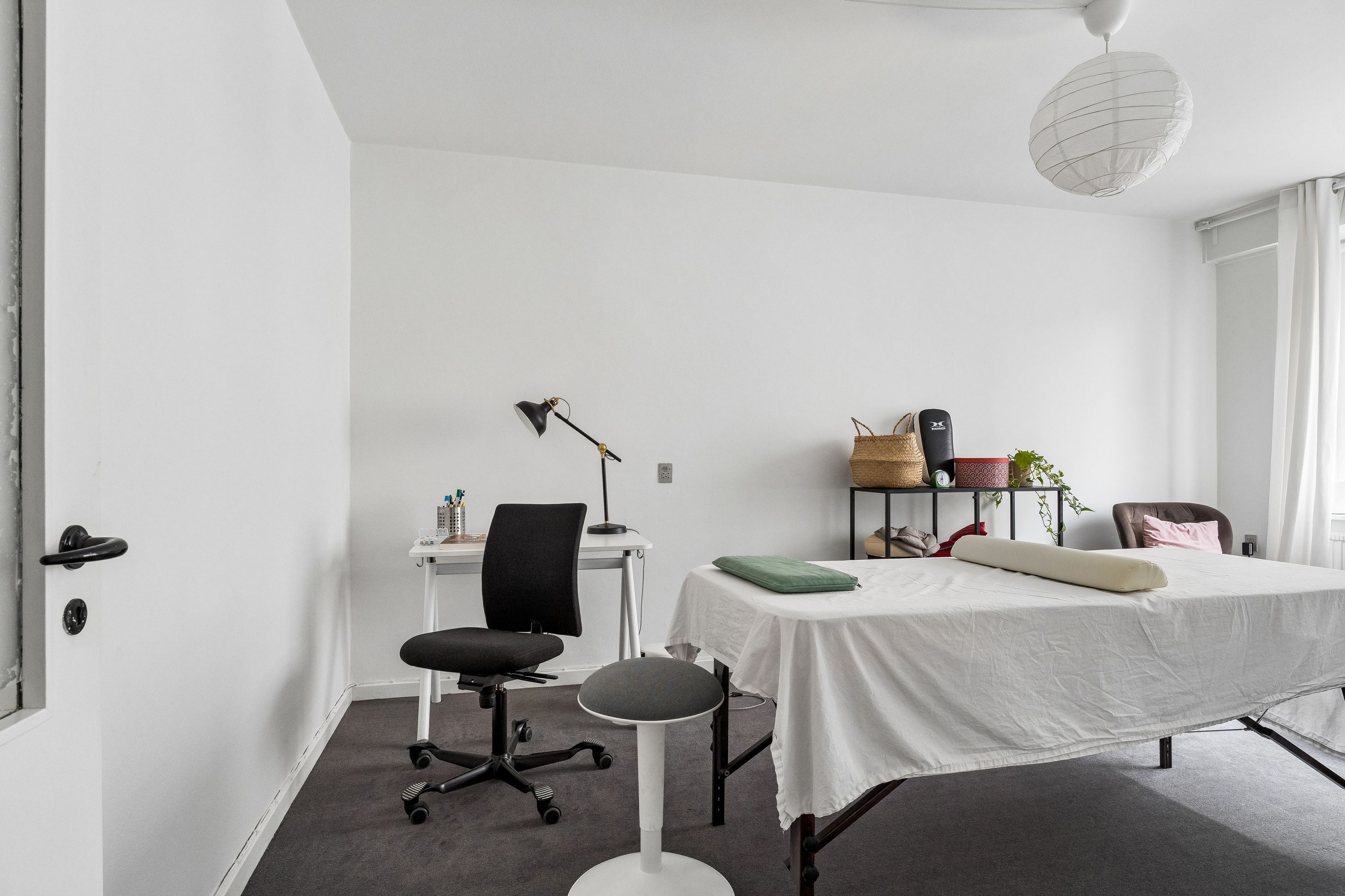 Kontor med højloft, hvidt bord, stol og lampe, massagebord med pude og hvid sengebetræk, to hængekøer og plante på hylde og lampeskærm og gardiner i hvidt og grønt