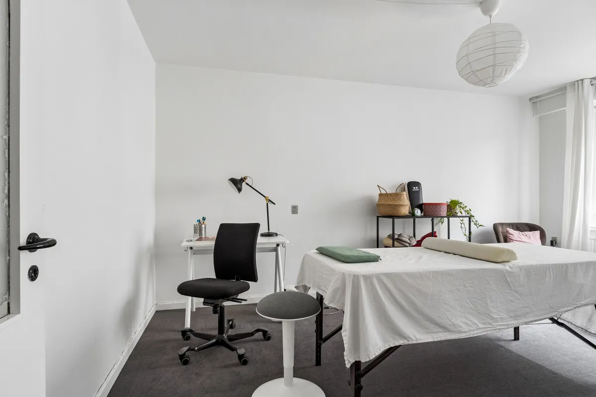 Kontor med højloft, hvidt bord, stol og lampe, massagebord med pude og hvid sengebetræk, to hængekøer og plante på hylde og lampeskærm og gardiner i hvidt og grønt