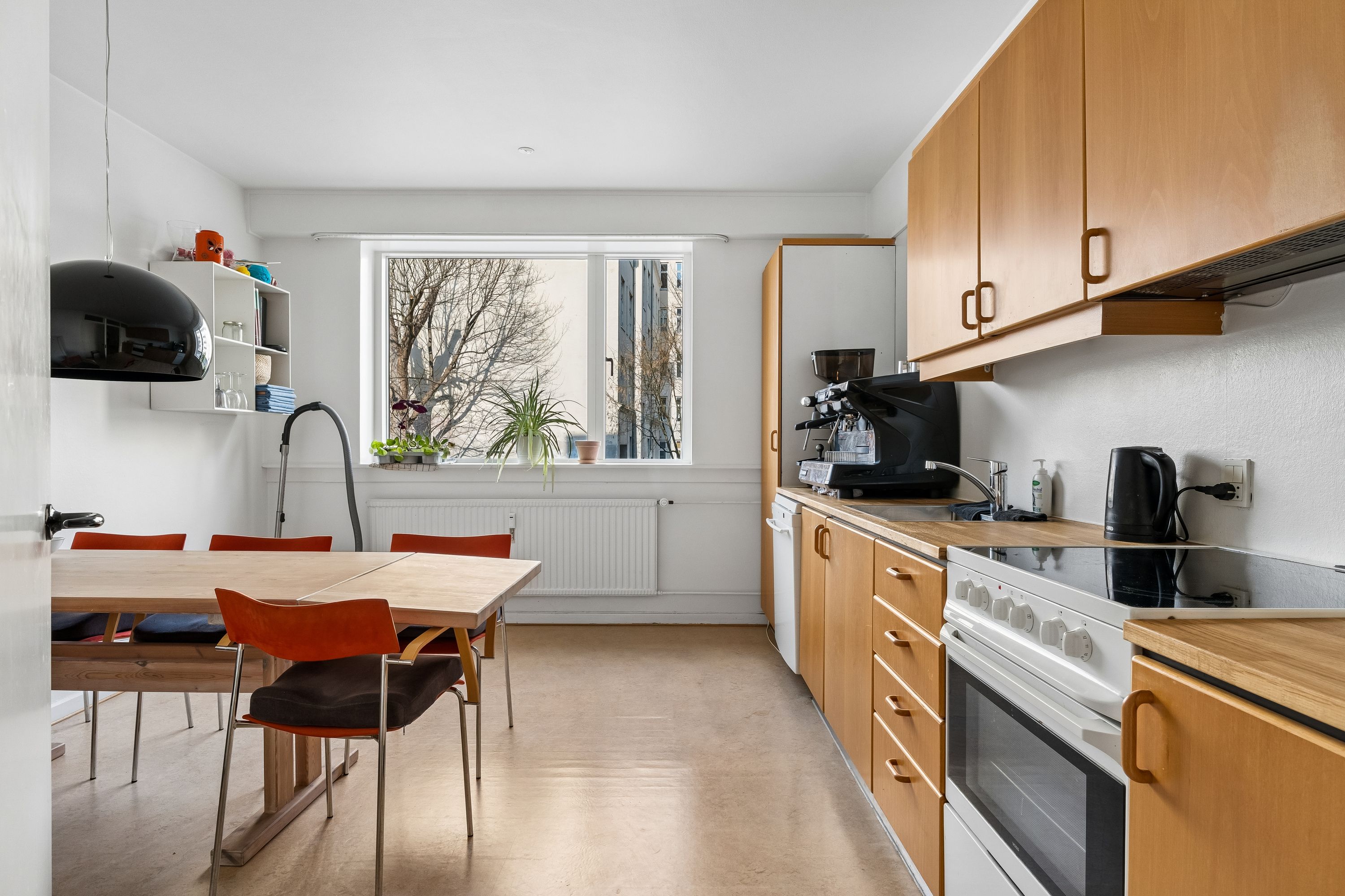 Moderne, minimalistisk køkken med hvide vægge, lysegrå gulv og lyse træskuffer. En hvid radiator og en stor vinduesflad med planter og et træbord med røde stole