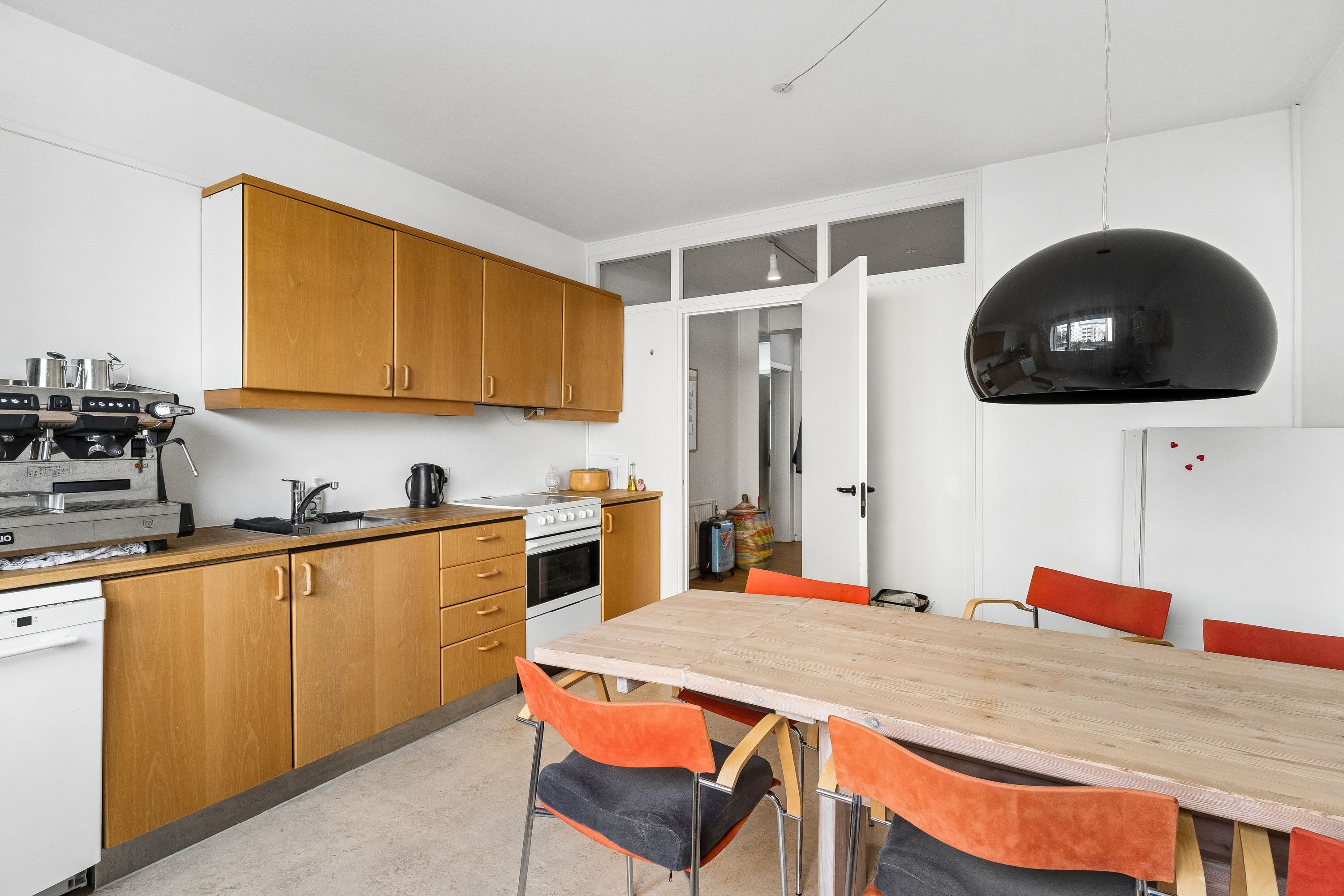 Moderne og funktionelt køkken med højloft, massivt træbord, orange stole og hvide loftsvægge, lysekroner og køkkenudstyr som en ovn og en kaffemaskine