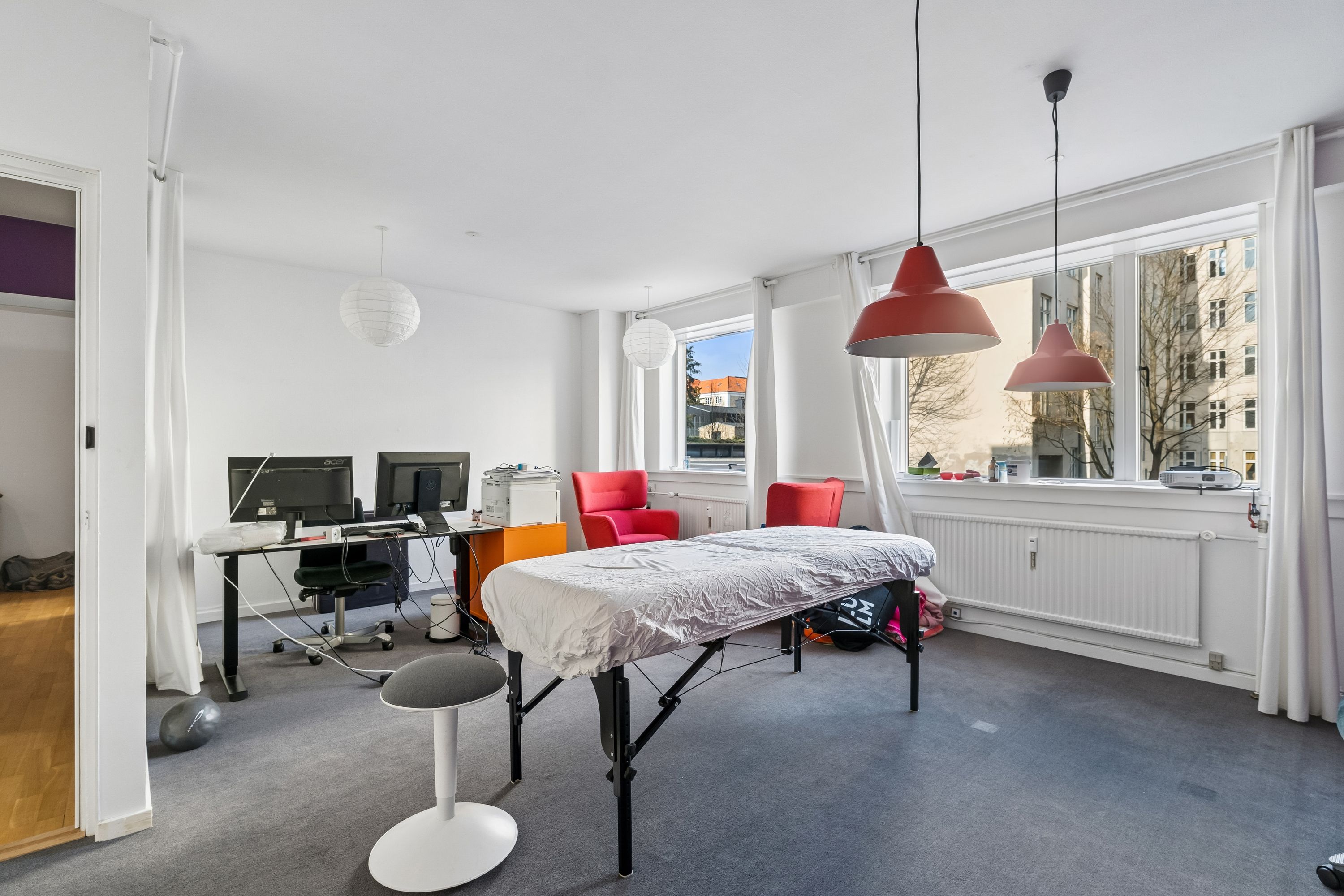 Kontor til leje med stort vindue, hvidt gulv, to skriveborde, to røde stole og to lysekroner. Et sengebord og en træningsklods er også synlige.