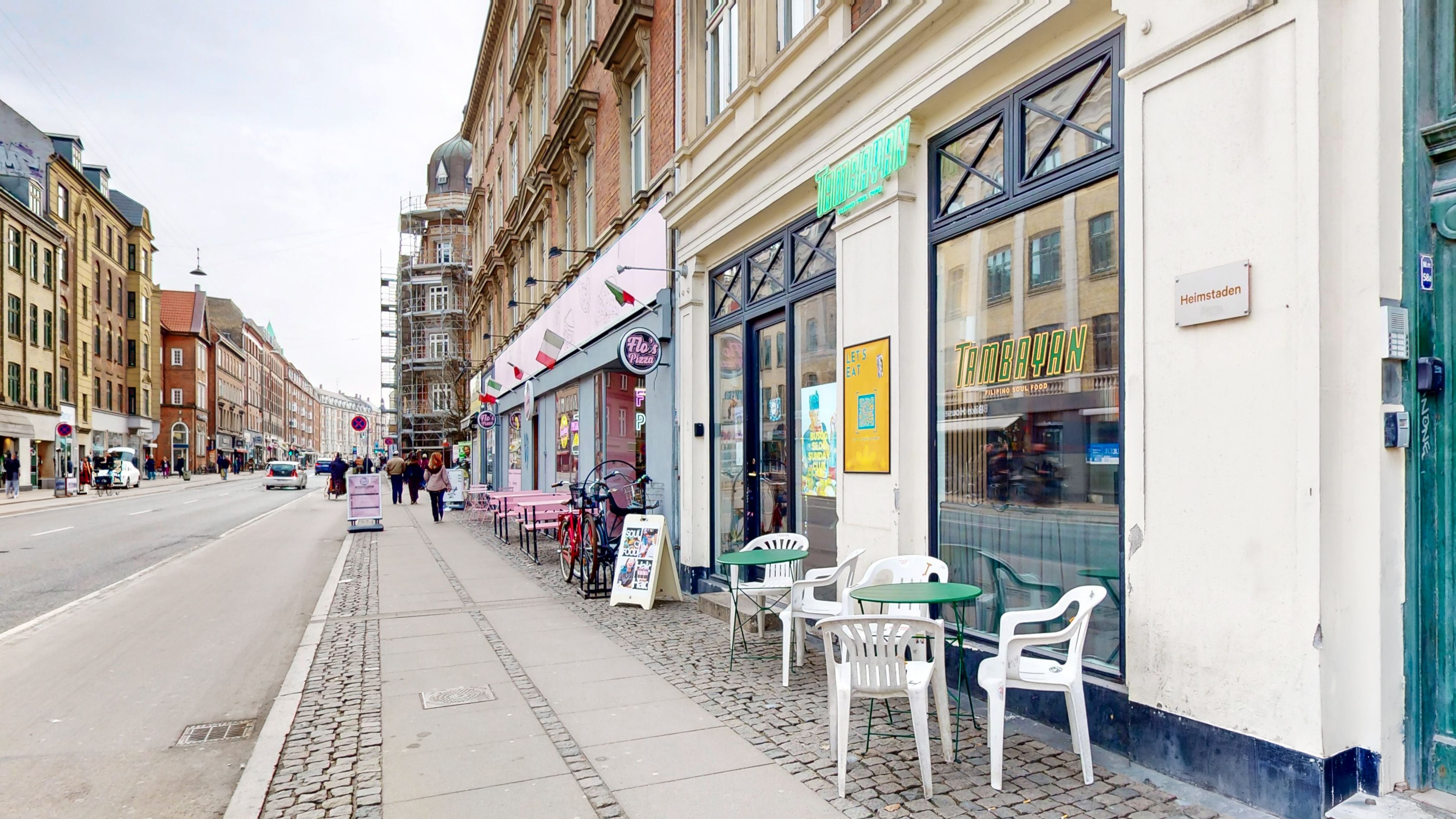 Bygning med restauranter og cafeer og cykler parkeret udenfor på Amagerbrogade. Der er flere folk, der går på gaden og kører biler.