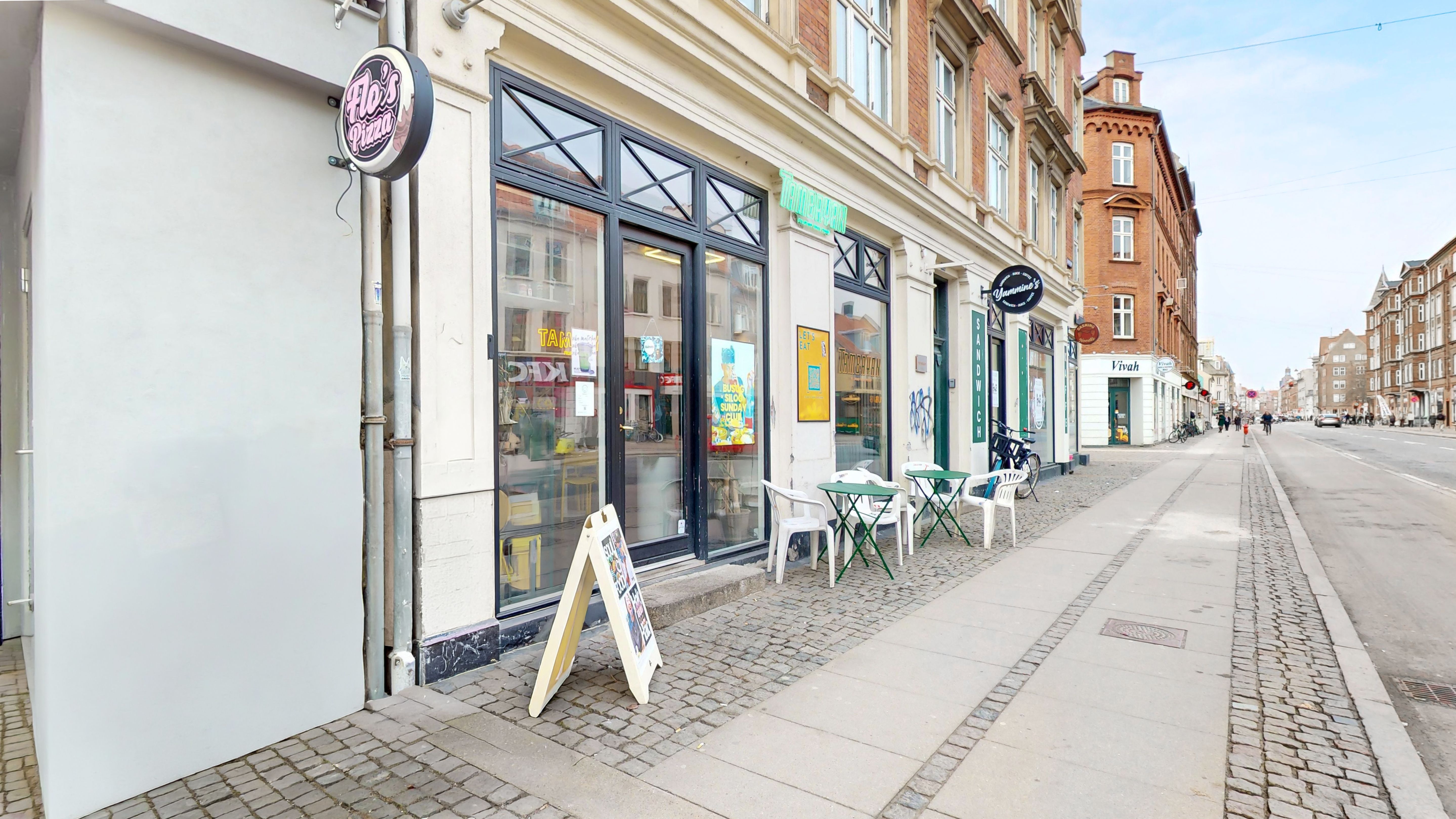 En pizzarestaurant og et sandwich-café med terrasseborde og stole på gågaden i København. Forretningen har glasruder og gul skrift.