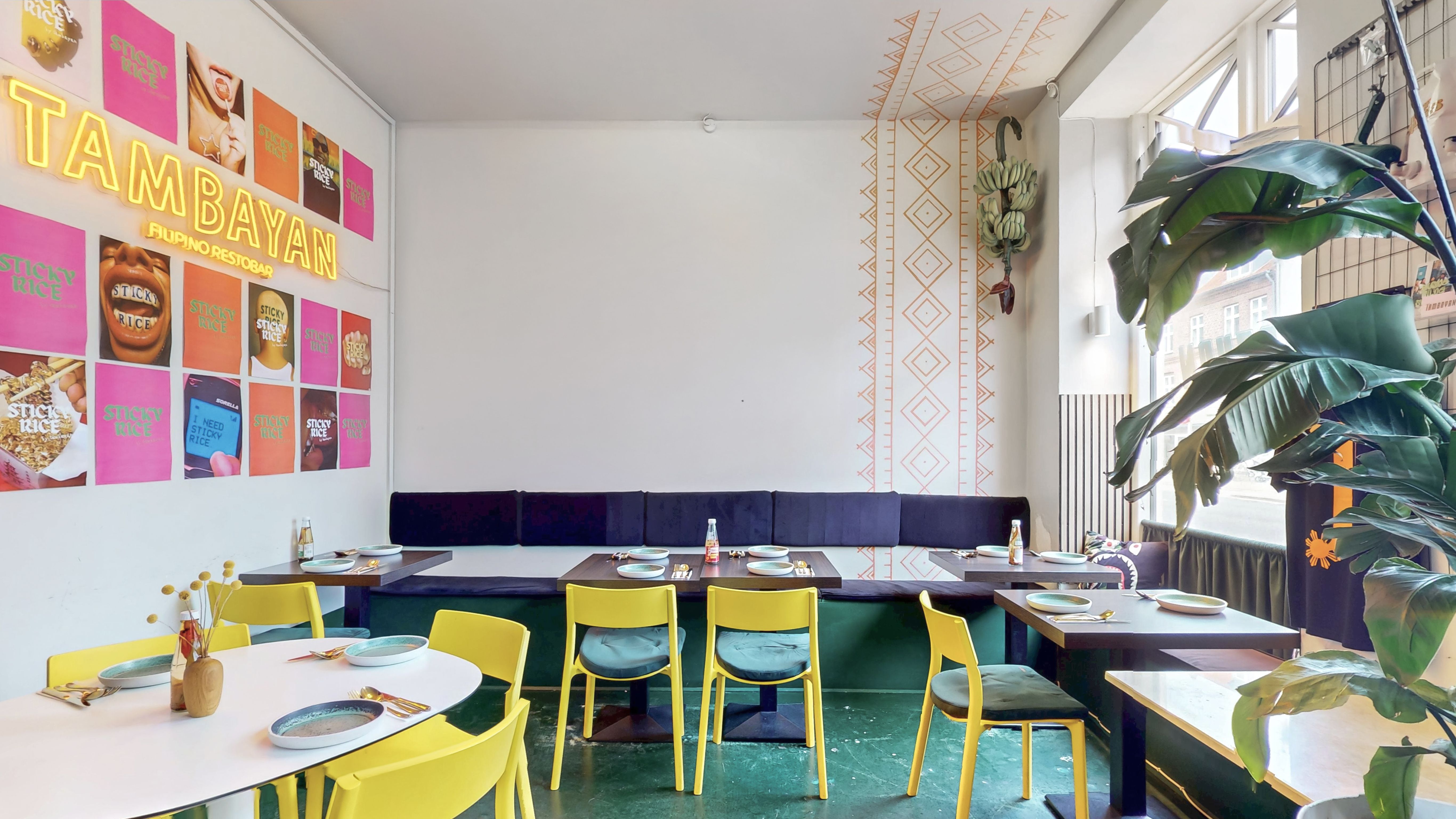 Væg med lyse, farverige plakater og gul neonskrift. Bord med plader og glas. Gule stole med blå pudsede sæder. Grønne planter og træer.