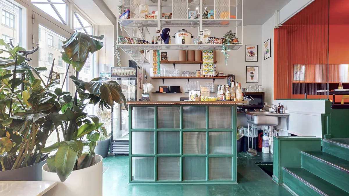 Selvbetjeningsrestaurant med grønne vægge, grøn gulv, glasvægge, plante i hvidt plantekar, lysekroner og en stor plante i en grå potte.