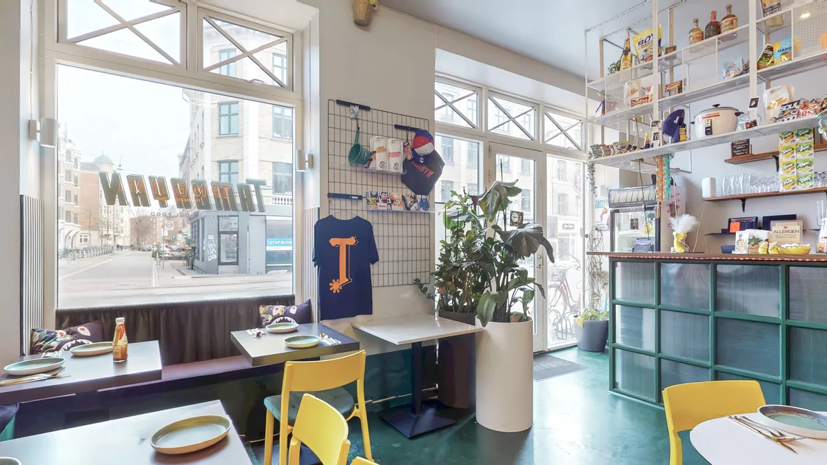 Inde i restauranten med lyse grønne vægge og gulv er der runde borde med gule stole. På bordene er der tallerkener og glas og der er blomster i en vase.
