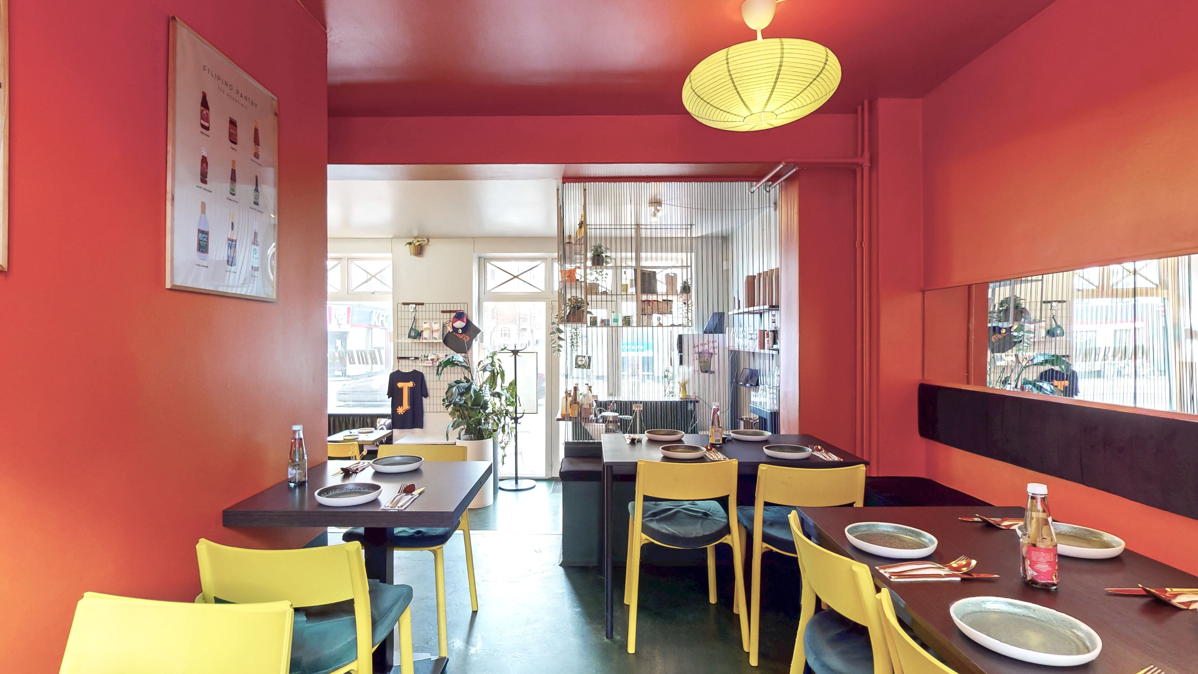 Inde i en restaurant med røde vægge, gule stole, to lange borde med serveret borde, en plante og en åbning til køkken.