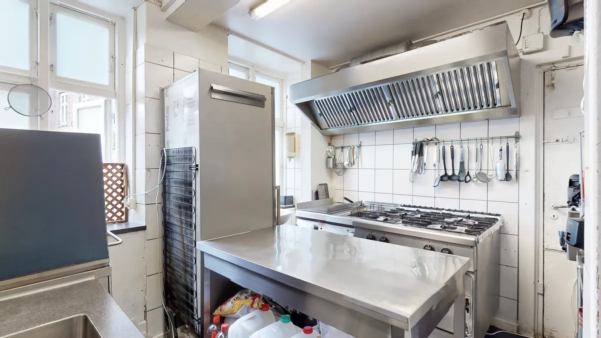 En kommerciel køkken med flere apparater, stålbord, ovn og gaskomfur, samt forskellige køkkenredskaber og ingredienser