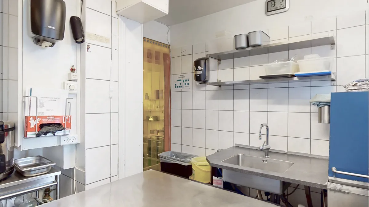 En professionel køkken med rustfrit stål vægoverbeslag og skabe, sæbeautomat og en enkelt vask, samt en hængende tørklædeholder og skraldspande.