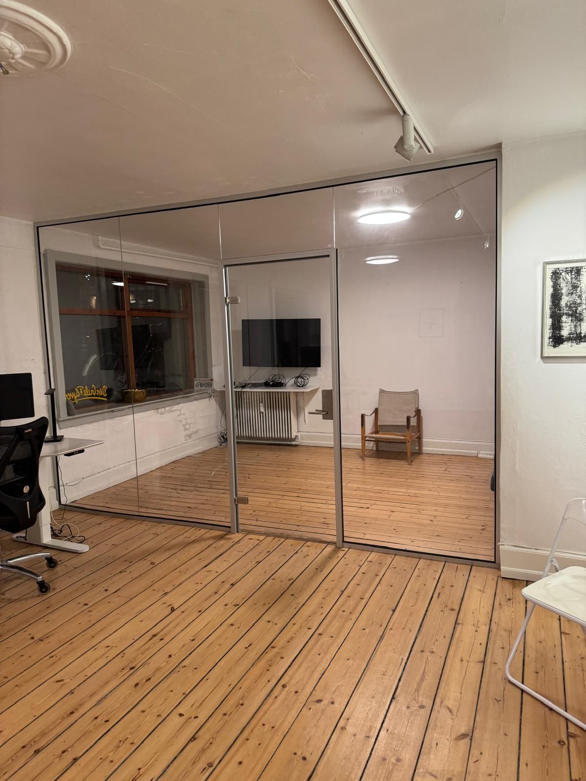 Kontor med glasvægge og trægulv. Rummet har en tv, en stol og et skrivebord. En lampe er installeret på loftet.