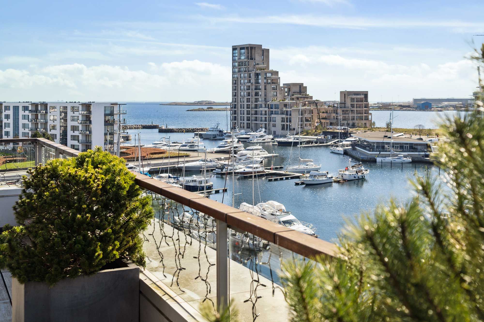 Havudsigt fra balkon med to små planter i gryder og udsigt over en stor havn med flere store og små både og bygninger