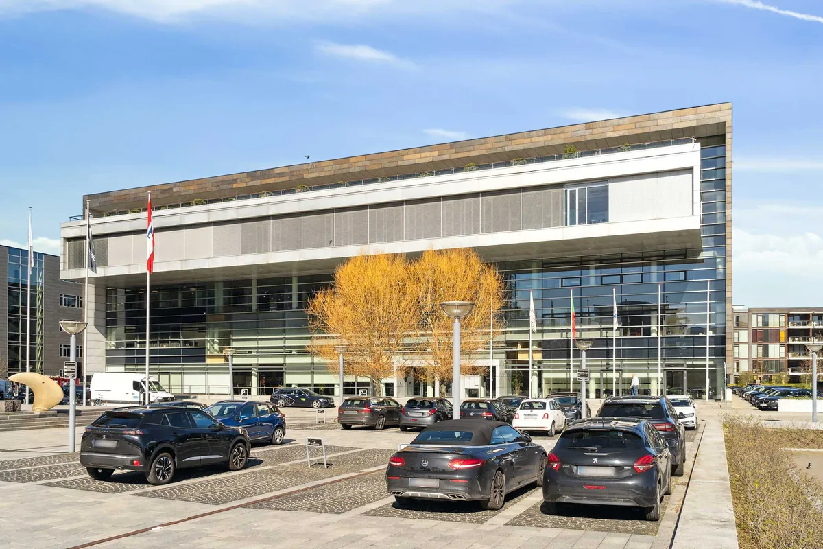 Markant bygning i lysebrun og hvid står foran parkeringsplads med parkerede biler og høje søjler og flag