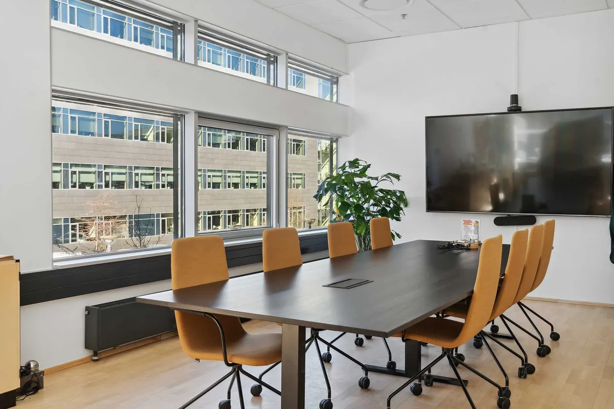 Moderne konferencelokale med glasvinduer, udstyr, plante og større tv. Bord og stol med hjul. Køkken med køleskab og stikkontakter.