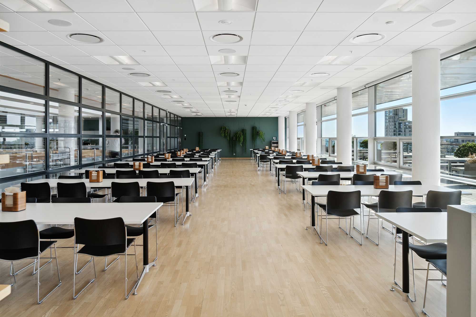 Et stort lysfyldt lokale med lysegult trægulv og lysegrå stolpe. En række borde og stole med tyk hvid overtræk. Glasser og planter i hjørner og vinduer med udsigt over byen